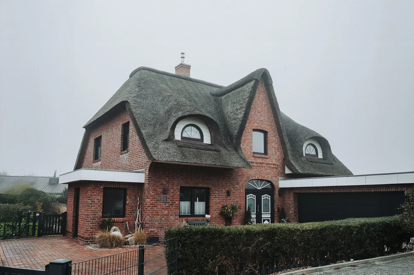 Mehrfamilienhaus mit altem, gekrümmtem Friesenhaus-Stil, rotem Backstein, friesisch gestaltetem Reetdach, schwarzen Türen und Fensterrahmen, umgeben von einem Garten und niedriger Hecke.