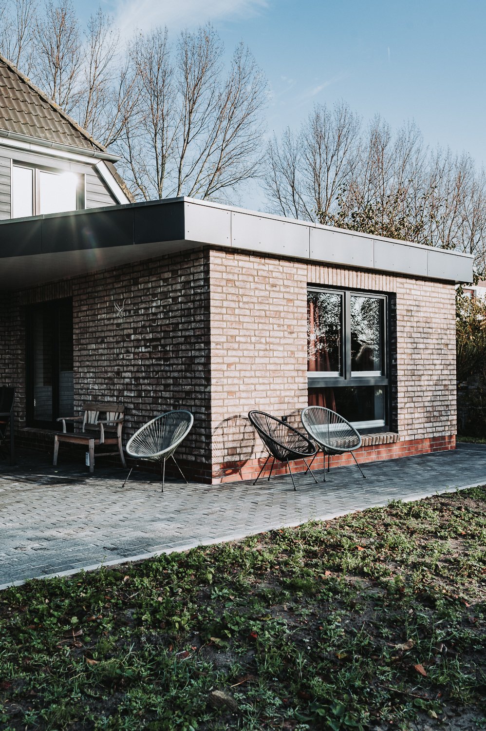 Ein modernes Haus mit einem braunen Backstein-Fassadenteil und einem schwarzen Fenster. Außerhalb des Hauses befinden sich einige Gartenmöbel, darunter ein Holzbank und vier Stühle aus Metall in Schwarz und Weiß. Der Boden ist mit Pflastersteinen aus
