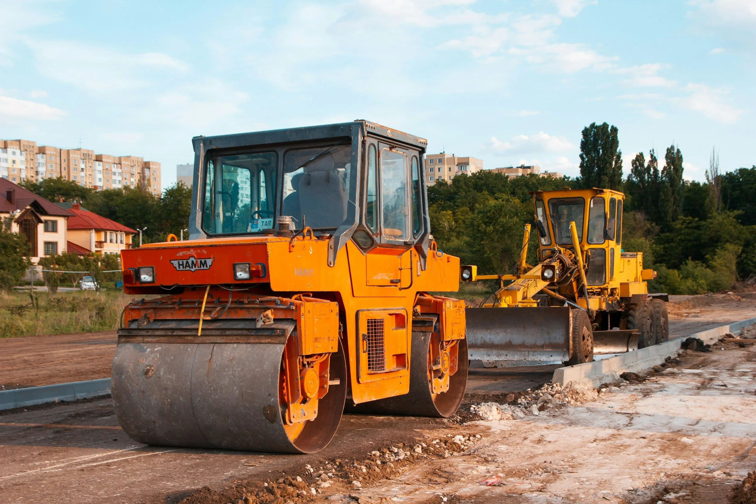 Zwei Baufräsen beim Wegbau auf einer Baustelle mit Wohnhäusern und Bäumen im Hintergrund.