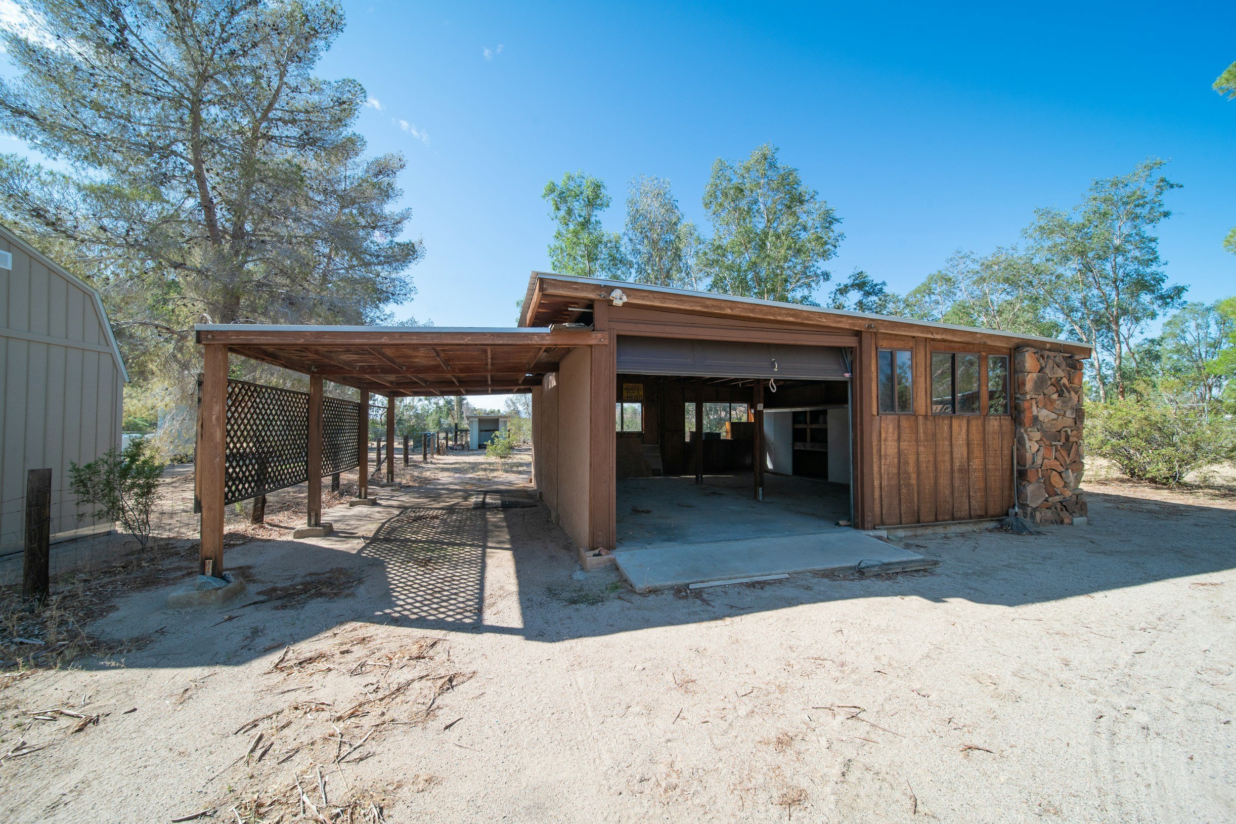 Ein offenes gemauerter und holzverkleideter Carport auf sandigem Boden, umgeben von Bäumen und einem blauen Himmel.