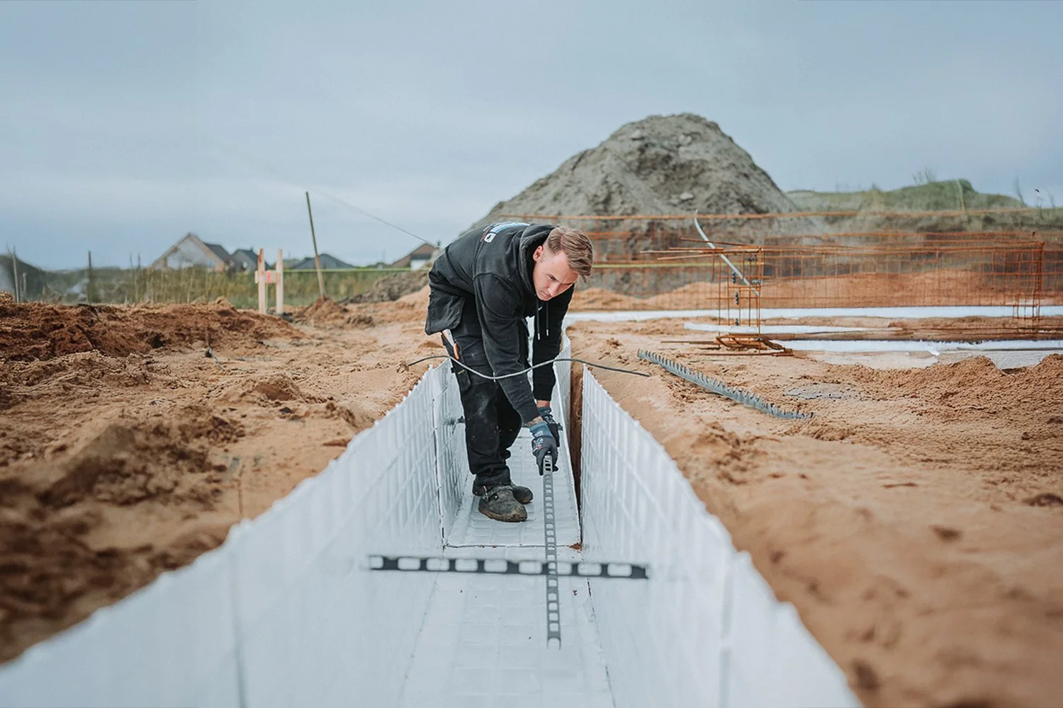 Ein Mann arbeitet auf einer Baustelle und legt ein betoniertes Fundament mit Kieselsteinen. Im Hintergrund sind Sandhügel und ein grauer Himmel.