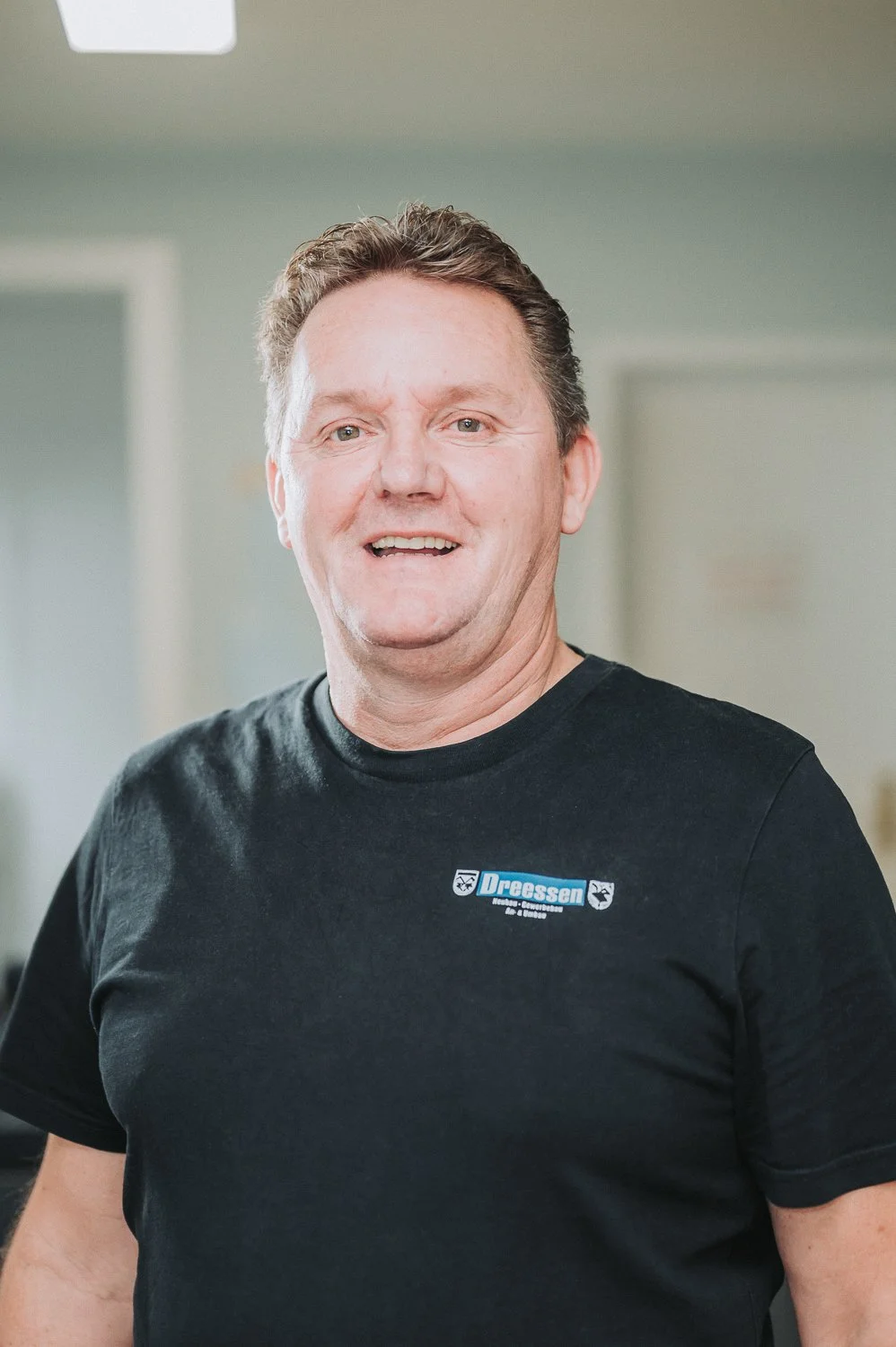 Ein lächelnder Mitarbeiter der Firma Dreessen Bau trägt ein schwarzes T-Shirt mit einem Logo und Text "Baugeschäft Dressen". Er steht in einem Raum mit hellen Wänden.