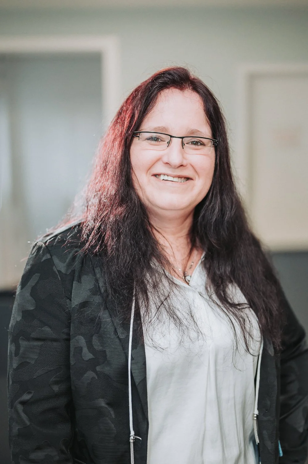 Eine Mitarbeiterin der Firma Dreessen  mit langen dunklen Haaren, Brille und einer schwarzen Jacke, lächelnd im Büro.