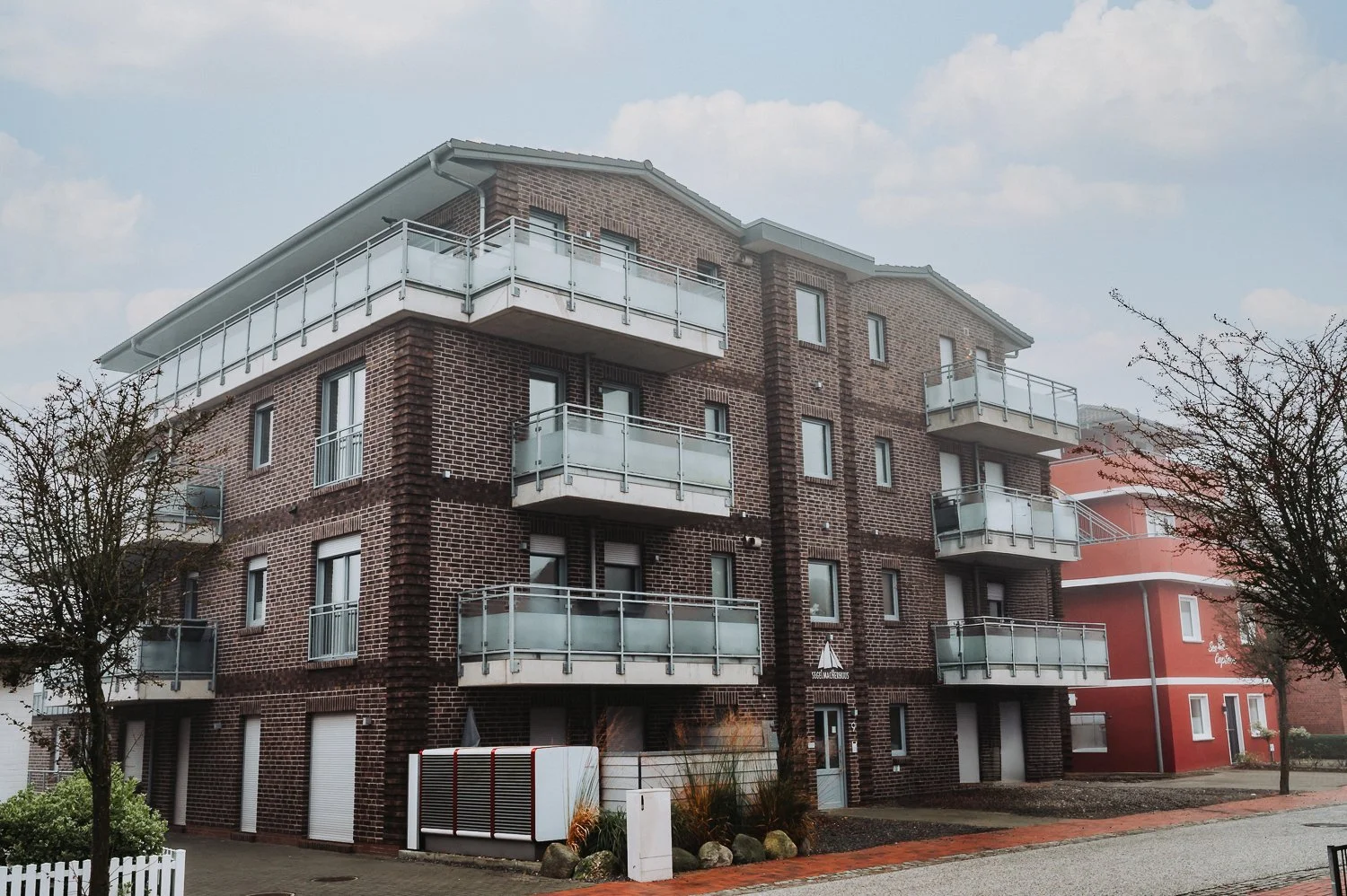 Mehrstöckiges Wohngebäude mit roten Ziegelwänden, weißen Balkonen und umgebenden Bäumen