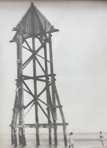 Ein Holz-Wasserturm am Strand mit zwei Kindern im Vordergrund.