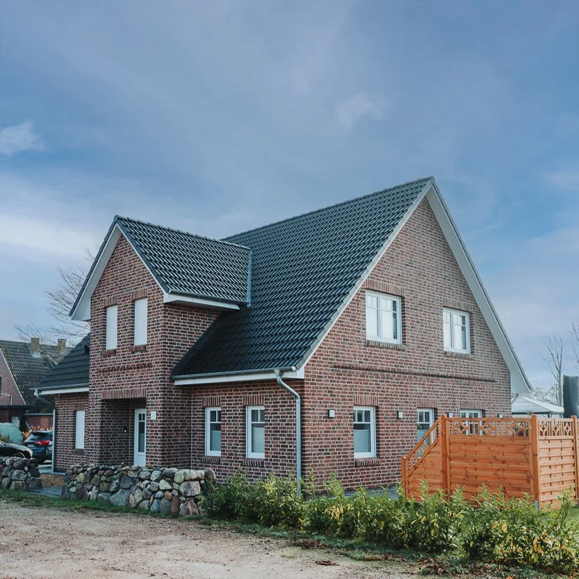 Ein zweistöckiges, rotes Backsteinhaus mit weißem Dach, mehreren Fenstern und einem kleinen Garten mit einer Holzsichtschutz.