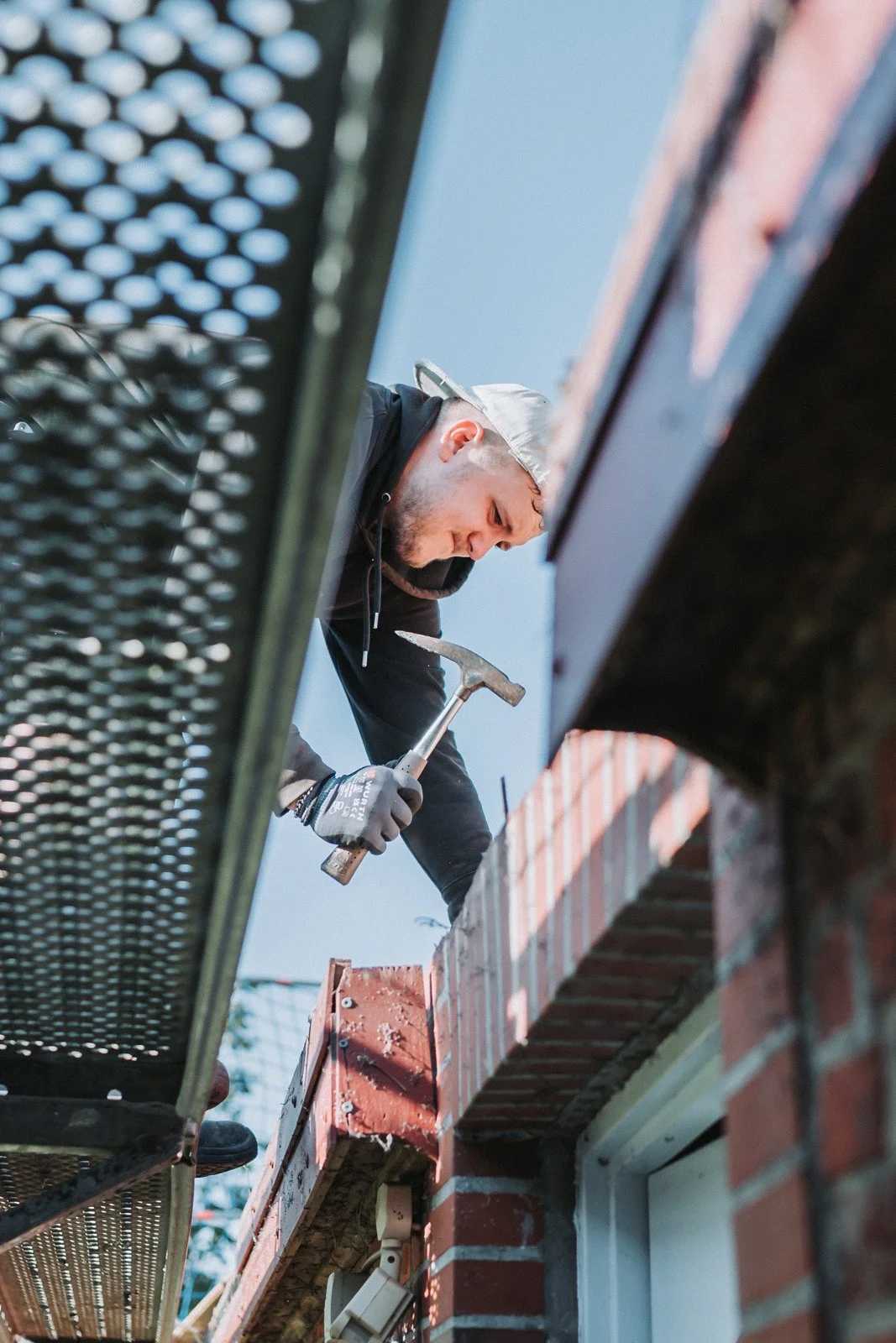 Ein Handwerker arbeitet auf einem Baugerüst an einem Haus, benutzt einen Hammer, trägt einen Helm und Handschuhe, um die Sicherheit zu gewährleisten.