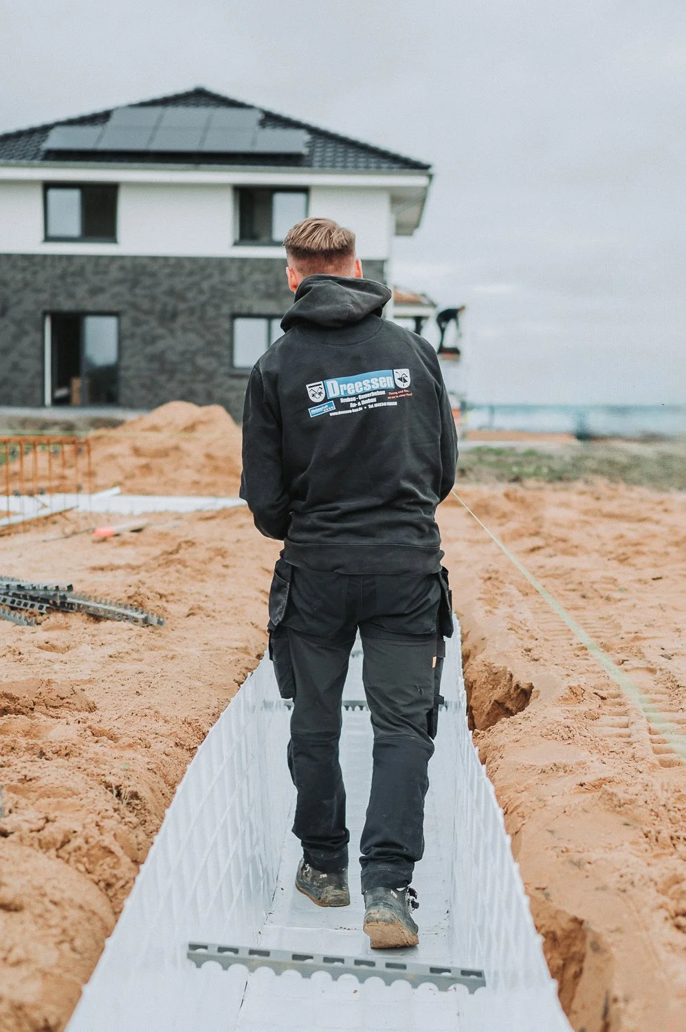 Eine Person trägt eine schwarze Jacke mit dem Logo 'Dreessen' und arbeitet an einem Baugrundstück, während sie auf einer weißen, isolierten Betonmatte geht. Im Hintergrund befinden sich ein modernes Haus und Bodenschüttung.