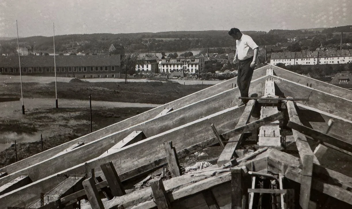 Schwarz-weiß-Foto einer Baustelle, auf der ein Mann auf einem Dach aus Holz arbeitet.
