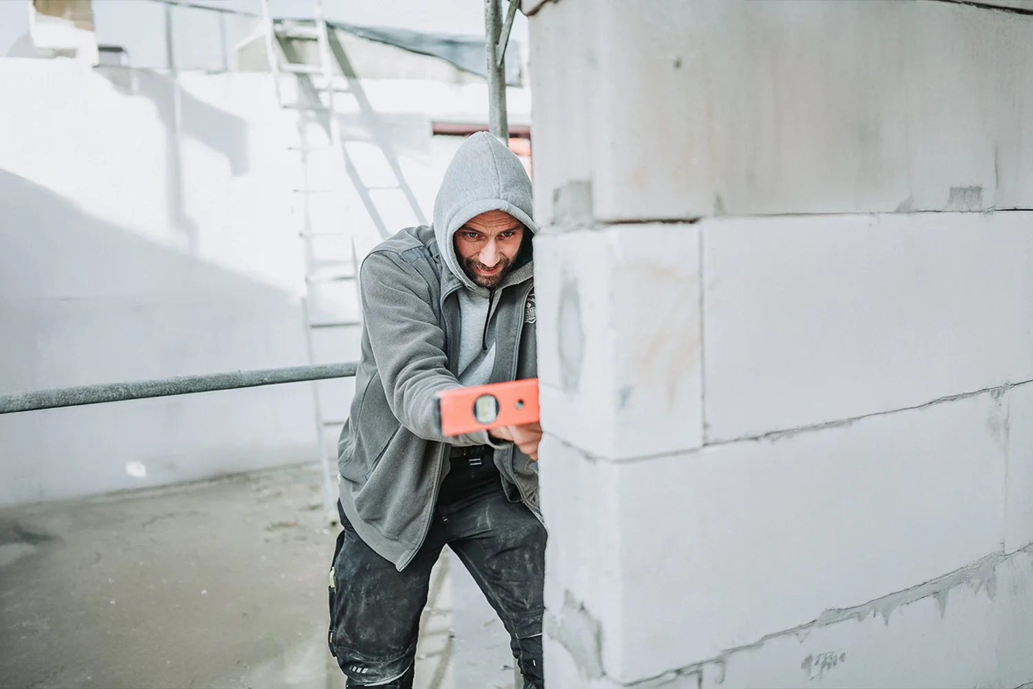 Mann in Kapuzenjacke und Arbeitshosen benutzt eine Wasserwaage, um die Wand auf einer Baustelle zu messen.