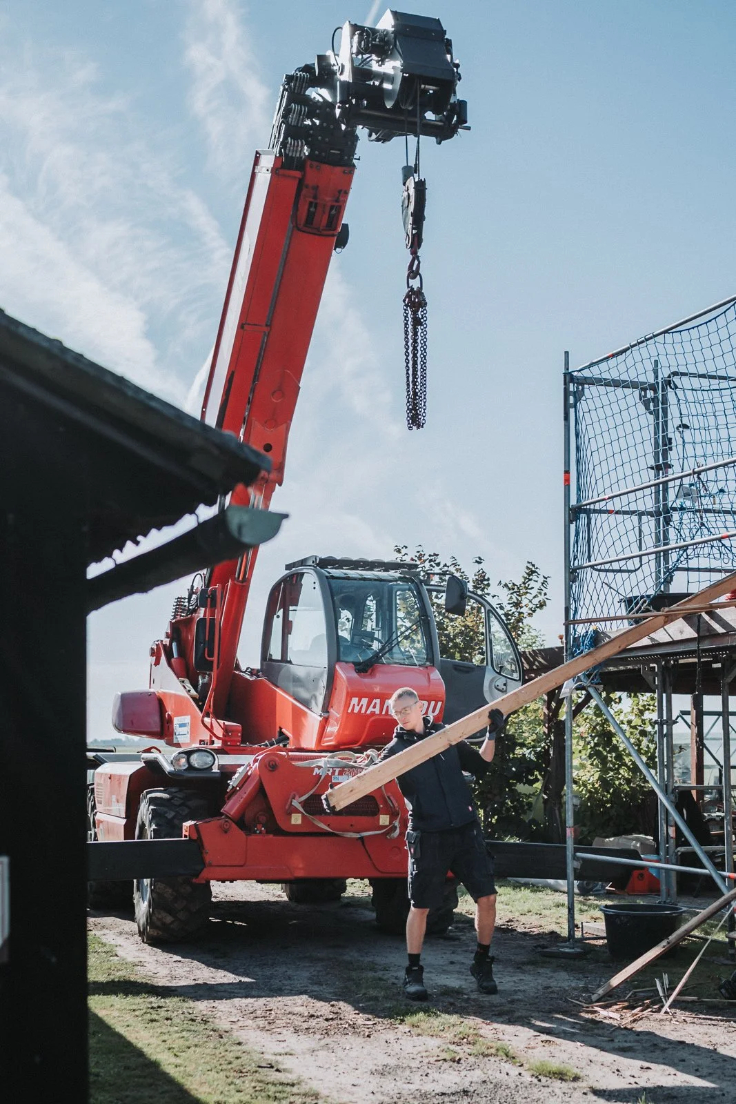 Ein Mann arbeitet auf einer Baustelle mit einem großen roten Kran. Der Kran trägt eine schwarze Hakenvorrichtung und befindet sich im Einsatz. Im Hintergrund sind Bäume und ein blauer Himmel mit einigen Wolken zu sehen.
