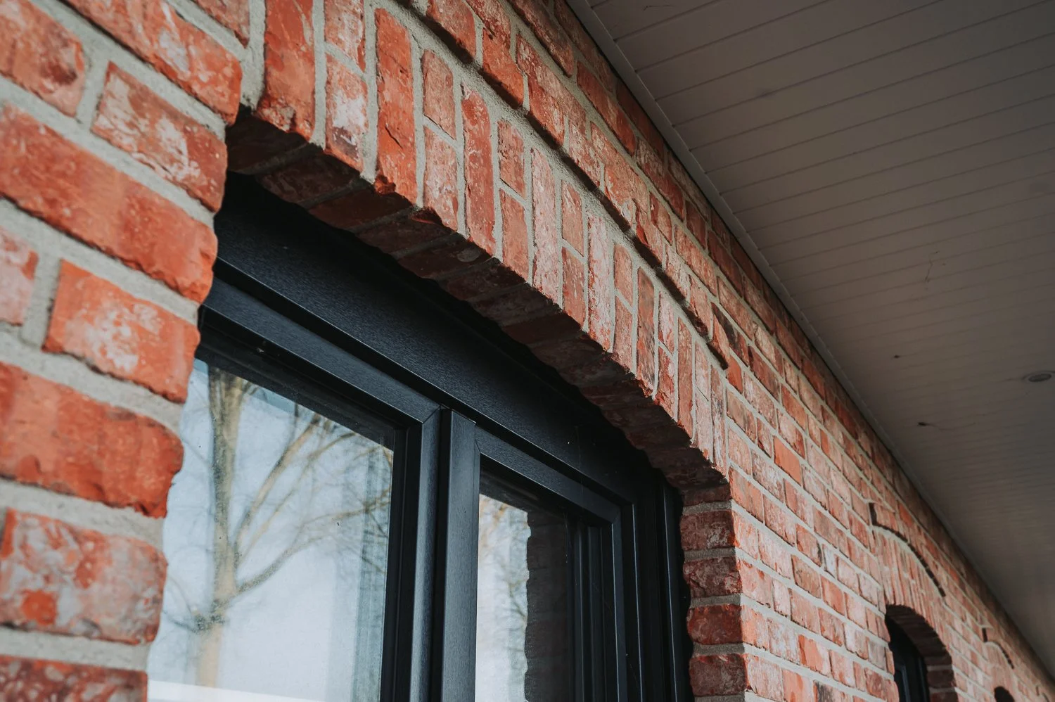 Außenansicht eines Gebäudes mit roten Ziegelwänden und schwarzen Fensterrahmen unter einem Vordach mit Holzverkleidung.