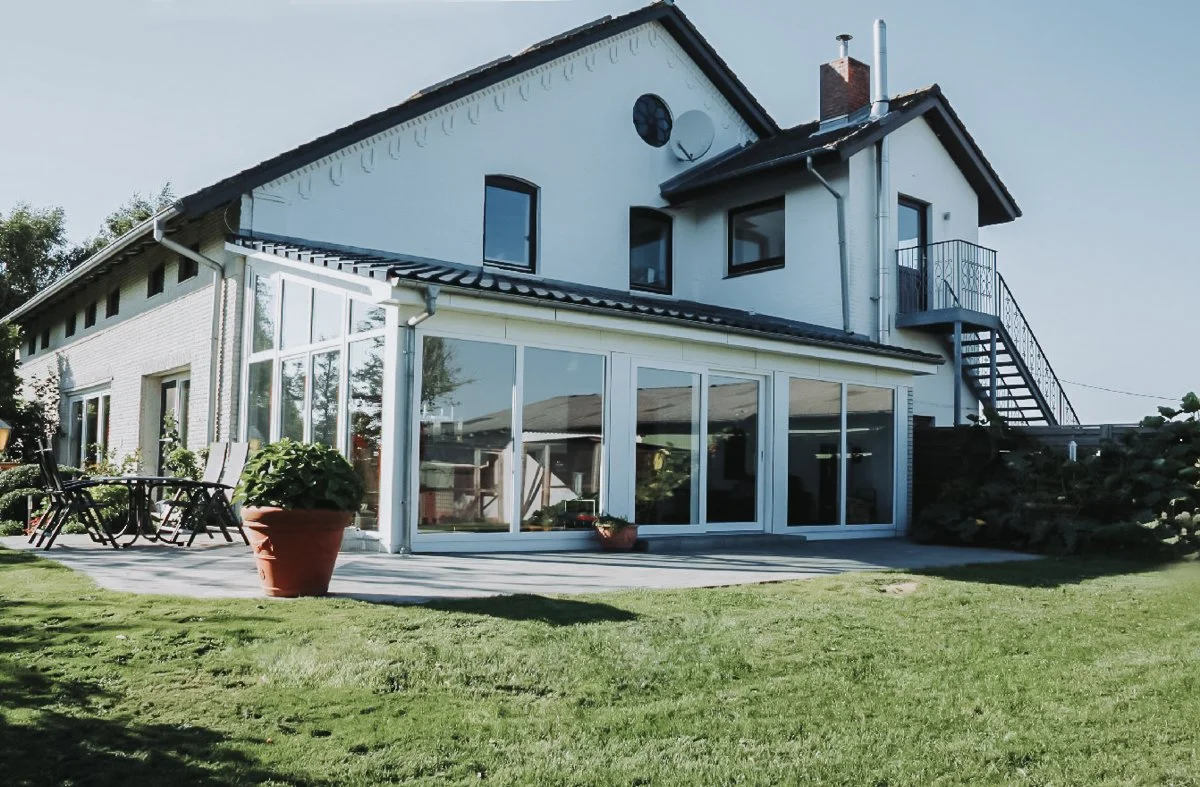 Ein zweistöckiges Haus mit weißen Wänden, großen Fenstern, einem Balkon, einer Treppe nach außen, einem gemähten Rasen und Gartenmöbeln im Vordergrund.
