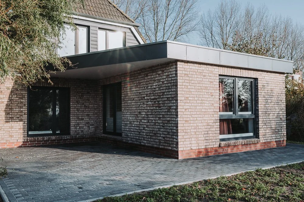 Modernes Haus mit Ziegelmauer, flachem Dach und großen Fenstern, umgeben von Bäumen.