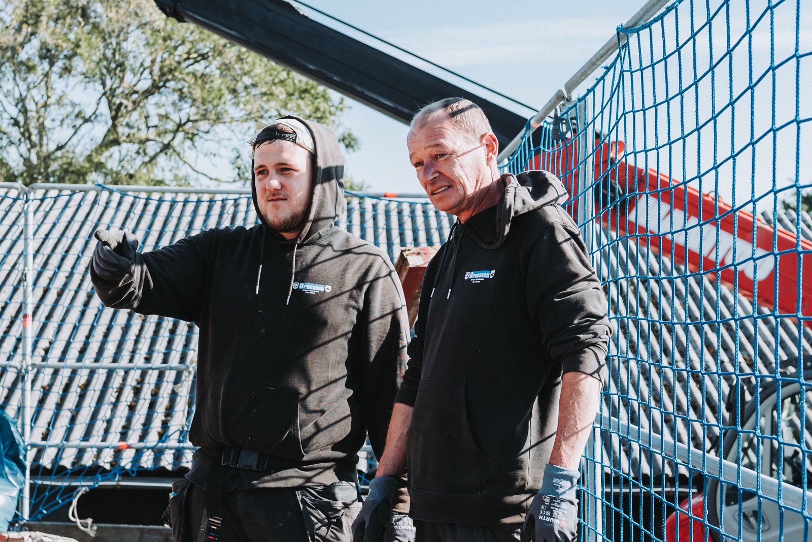 Zwei Männer in schwarzen Hoodies stehen neben einem blauen Sicherheitstreppengeländer. Der jüngere Mann zeigt nach links, während der ältere Mann mit ernstem Gesichtsausdruck den Blick nach vorne richtet.
