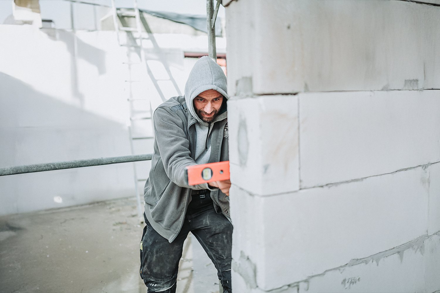 Ein Mann im grauen Kapuzenpulli und schwarzen Hosen überprüft ein Mauerwerk mit einer Wasserwaage auf einer Baustelle.