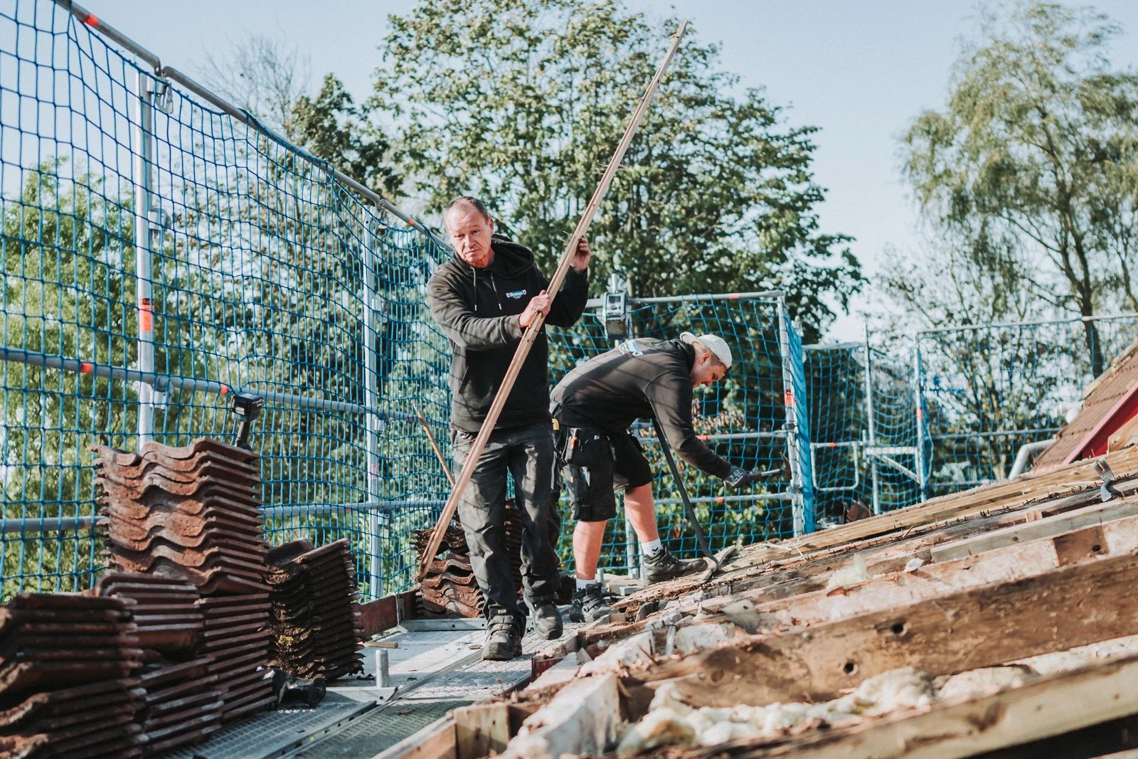 Zwei Männer beim Dachdeckerarbeiten auf einem Dach mit Dachziegeln, im Hintergrund Bäume und ein blauer Himmel.