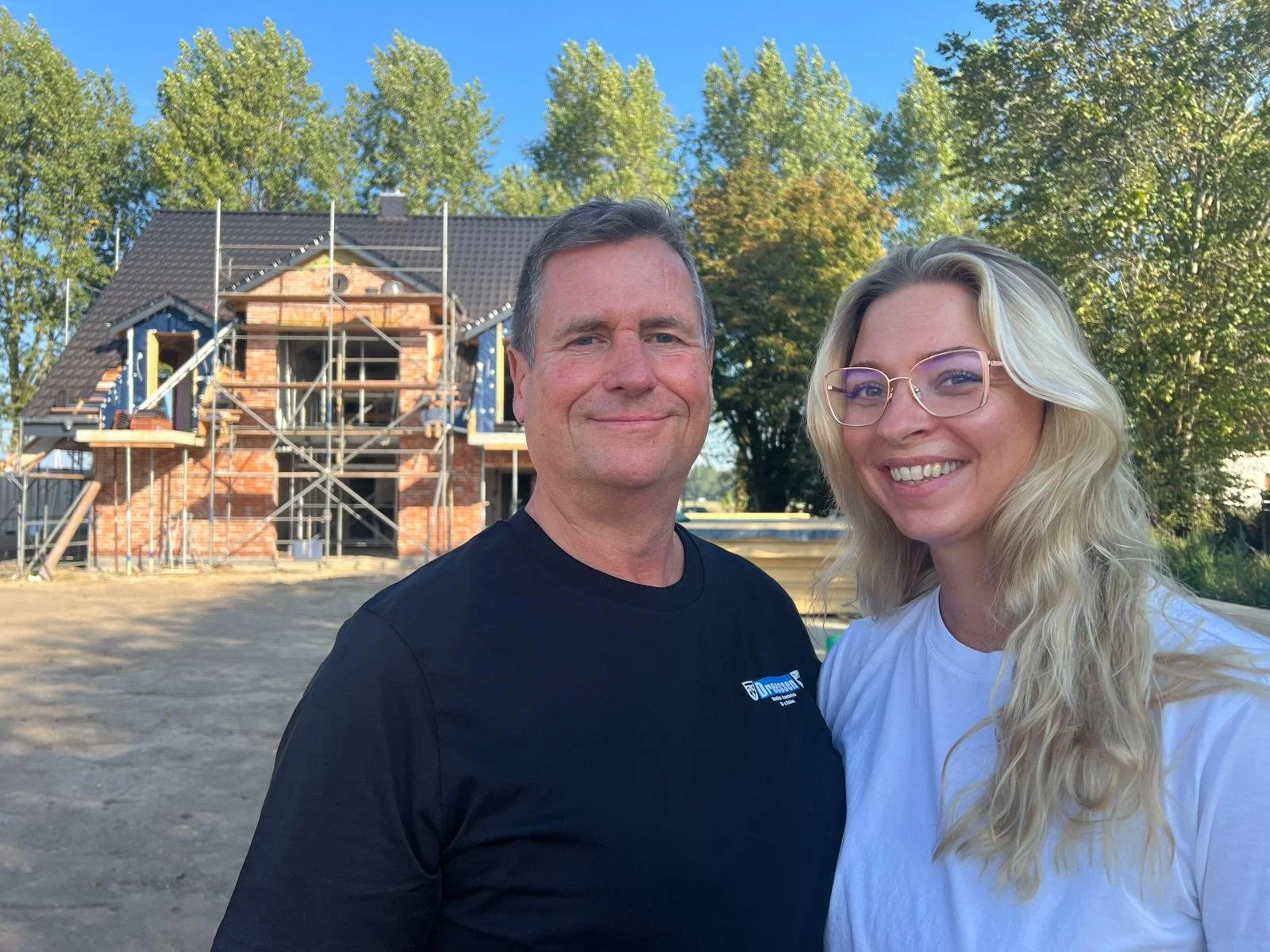 Ein Mann und eine Frau stehen vor einem unter Bau befindlichen Haus, umgeben von Baugerüsten und Bäumen bei sonnigem Wetter.