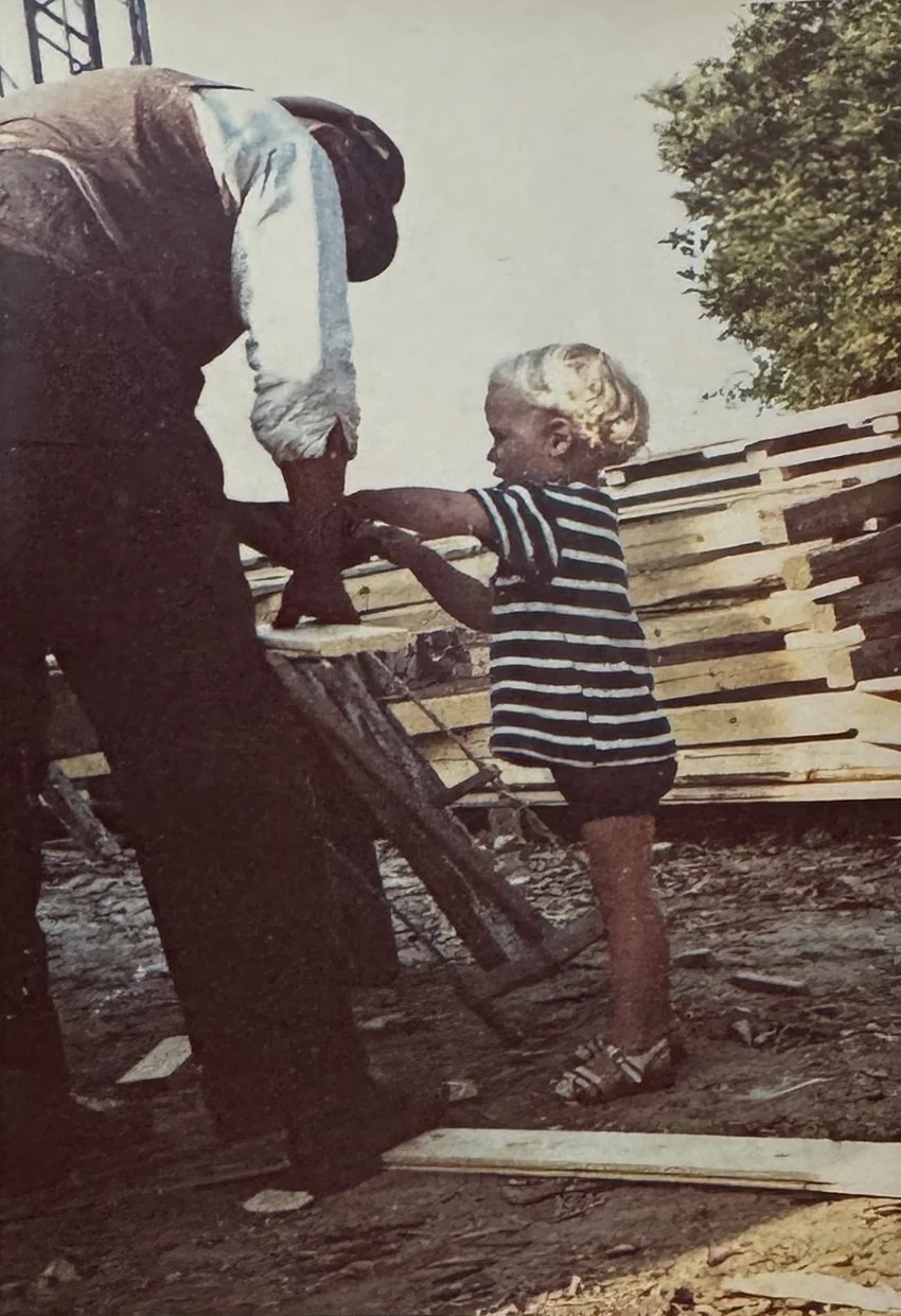 Ein Kind mit kurzen, blonden Haaren trägt ein schwarz-weiß gestreiftes T-Shirt und Shorts und reicht einem Erwachsenen einen Hammer bei einer Holzarbeit. Der Erwachsene trägt dunkle Kleidung und einen Hut. Es sind Holzbohlen gestapelt im Hintergrund,