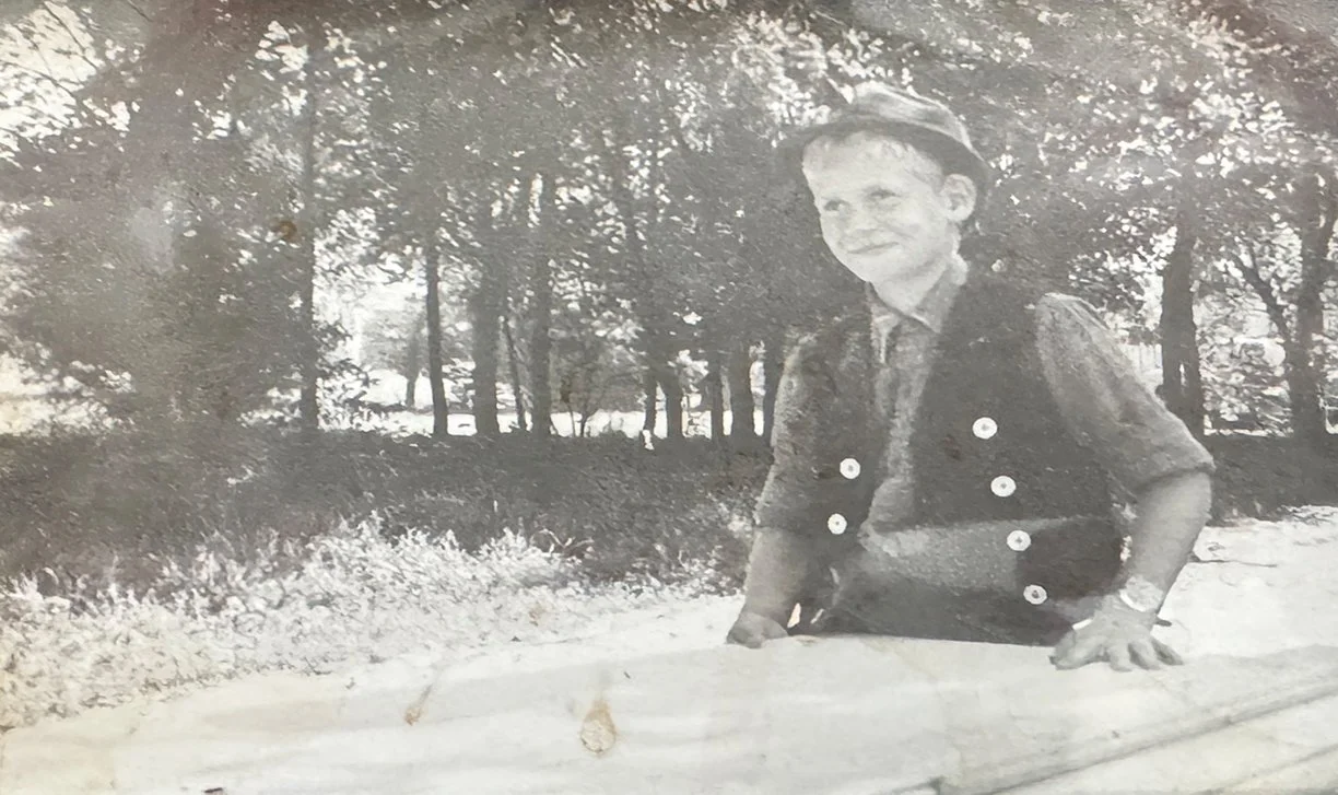 Schwarz-Weiß-Foto eines lachenden Jungen, der auf einem Baumstamm im Park sitzt, mit Bäumen im Hintergrund.