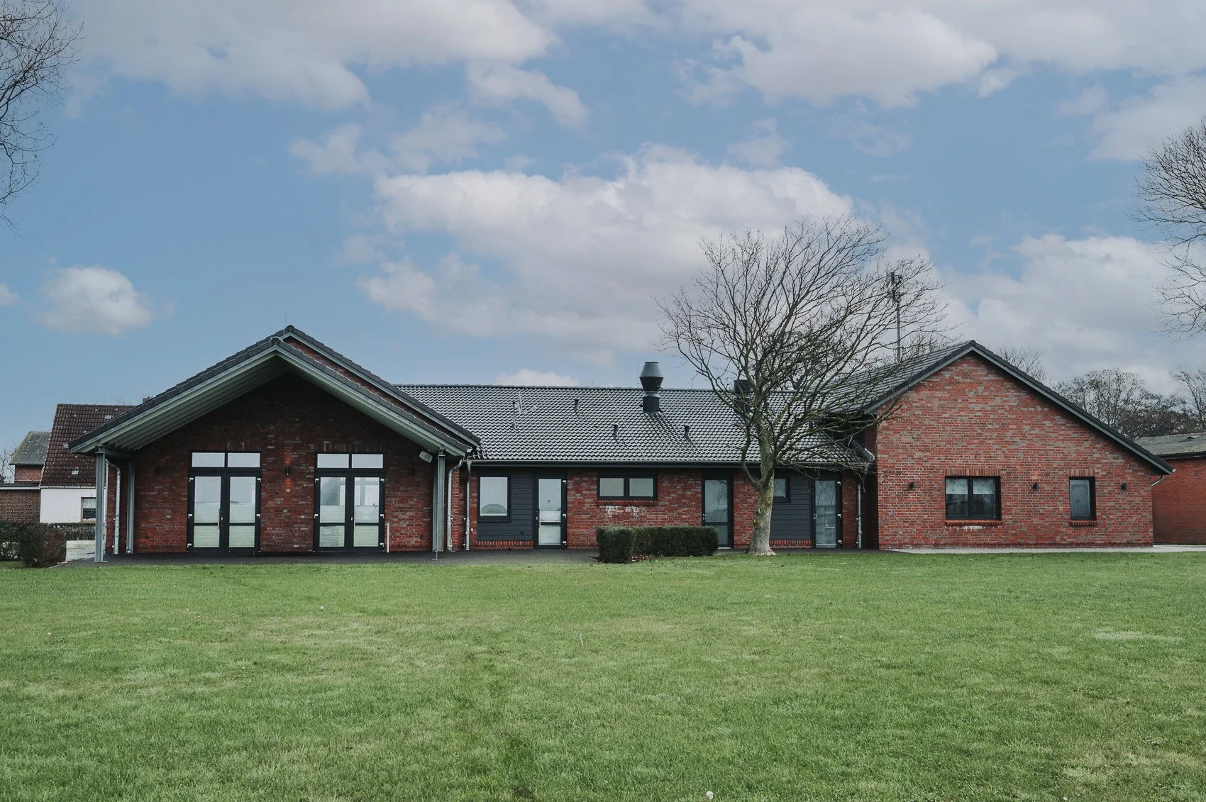 Ein modern gebautes Haus mit roten Backsteinwänden, schwarzen Fenstern und einem grauen Dach, umliegender grüner Rasen und einige Bäume, blauer Himmel mit Wolken.