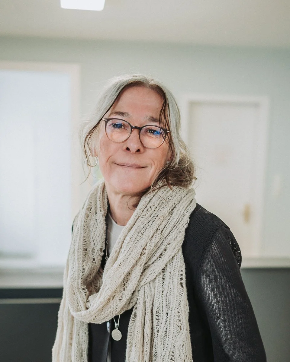 Eine ältere Frau mit Kurzhaarfrisur, Brille und einem weißen Strickschal um den Hals, trägt ein schwarzes Oberteil und steht in einem hellen Raum.
