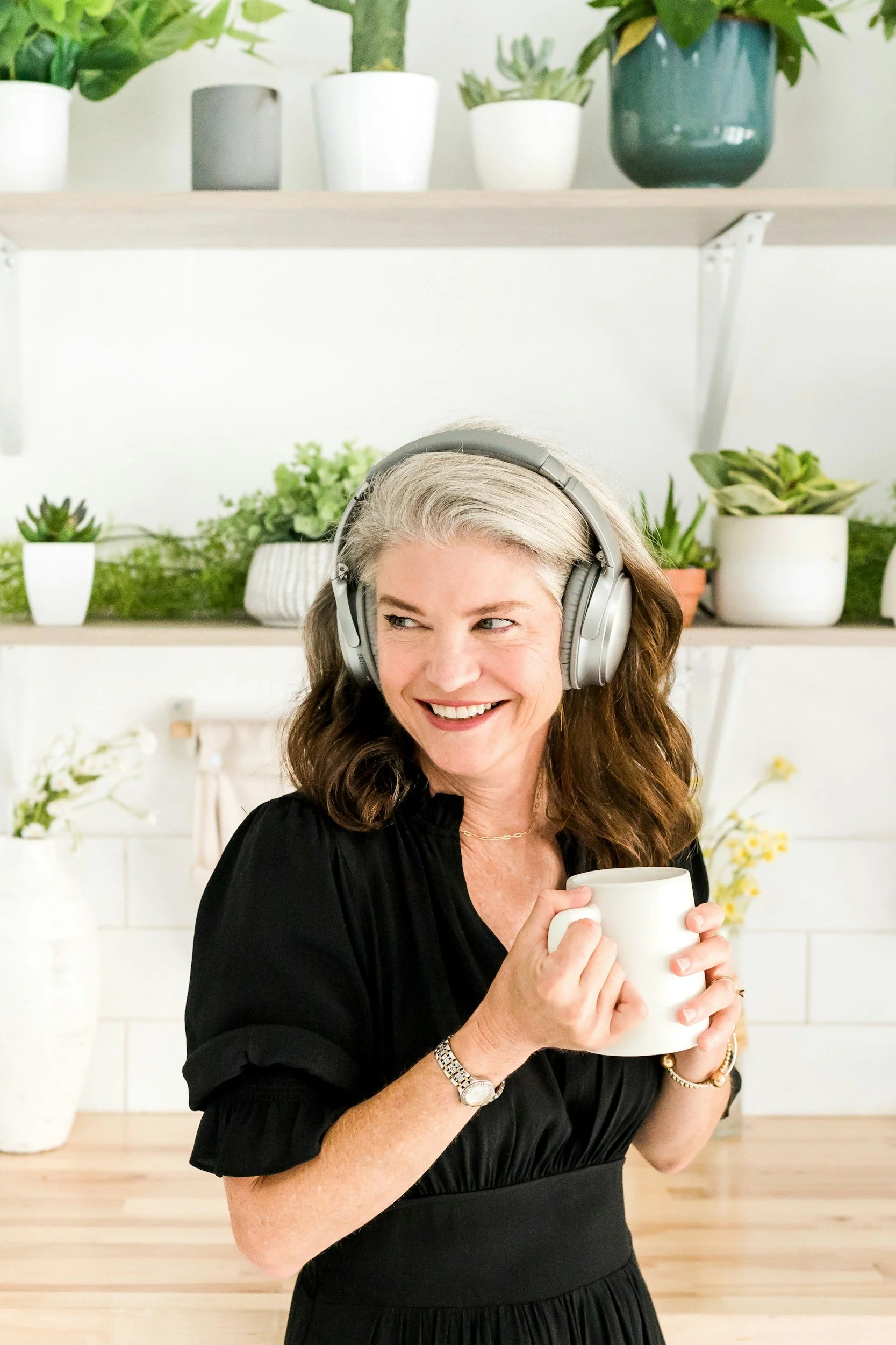 Smiling woman with gray hair, wearing headphones and a black dress, holding a white mug, in a room with shelves of potted plants.