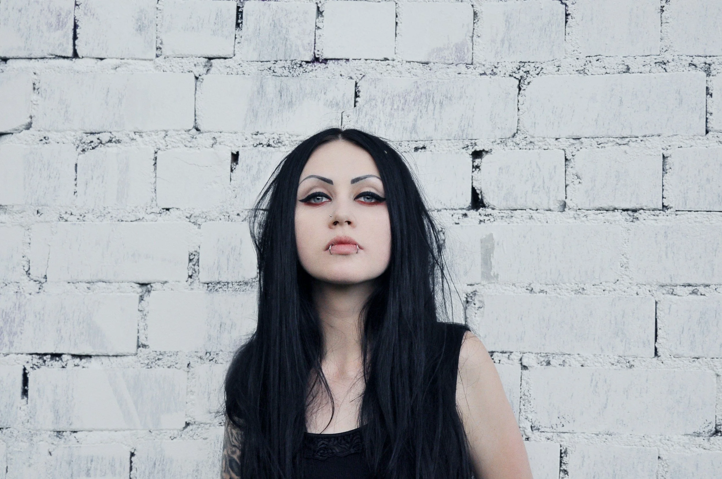 A young woman with long black hair, pale skin, and dramatic makeup standing against a white brick wall. She has piercings on her lips and nose and wears a sleeveless black top.