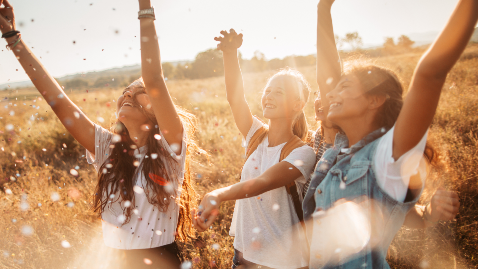 Group of four young women outdoors celebrating, throwing confetti, smiling, and raising their hands in the air during sunset.