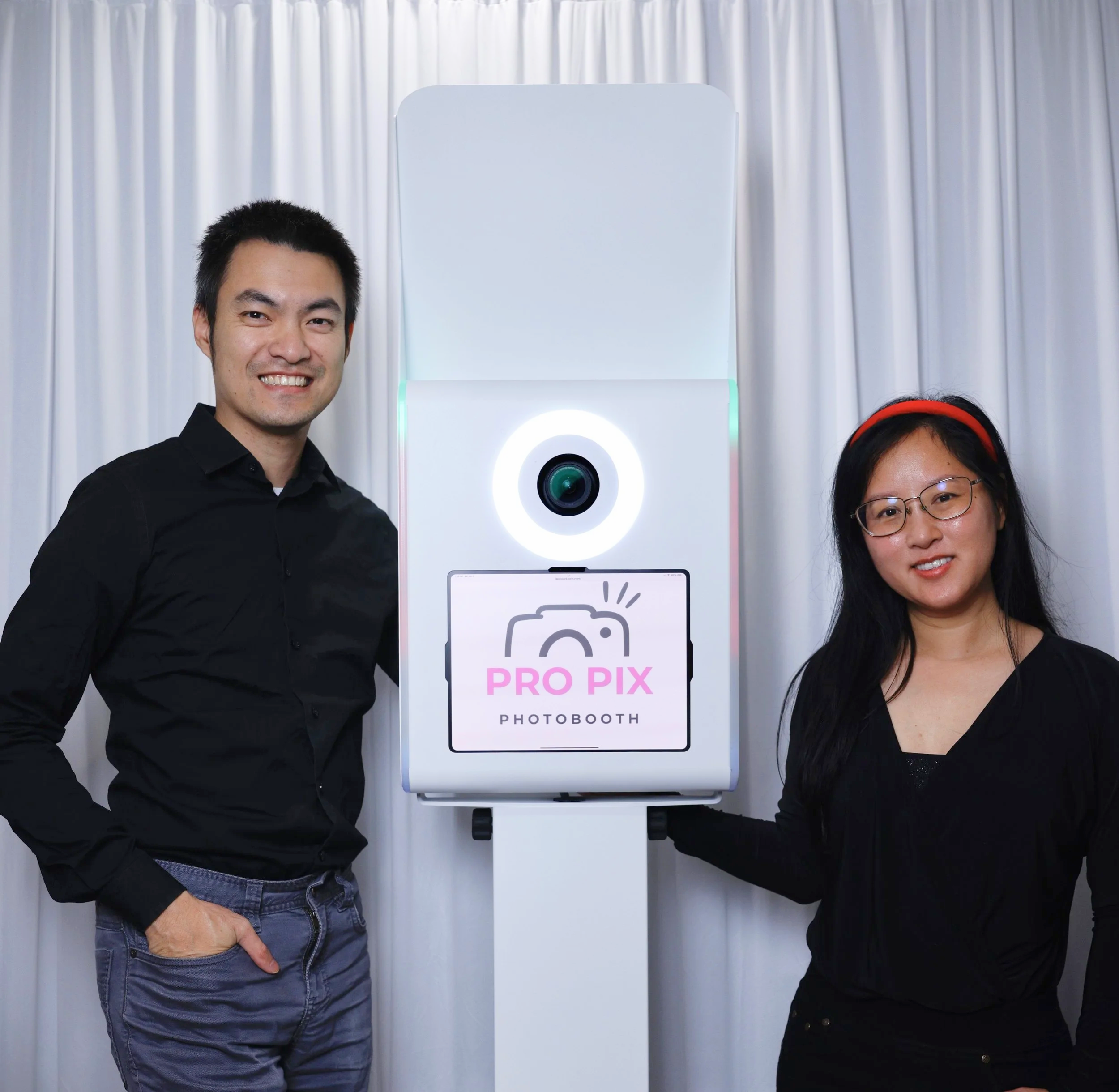 Two people, a man and a woman, stand on either side of a photo booth labeled 'PRO PIX PHOTOBOTH' with a camera icon, in front of a white curtain.