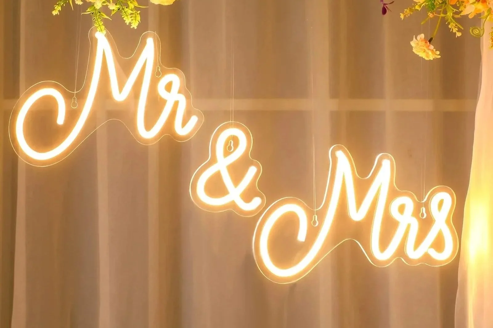 Neon sign reads 'Mr & Mrs' on a beige curtain background, with some greenery and flowers overhead.