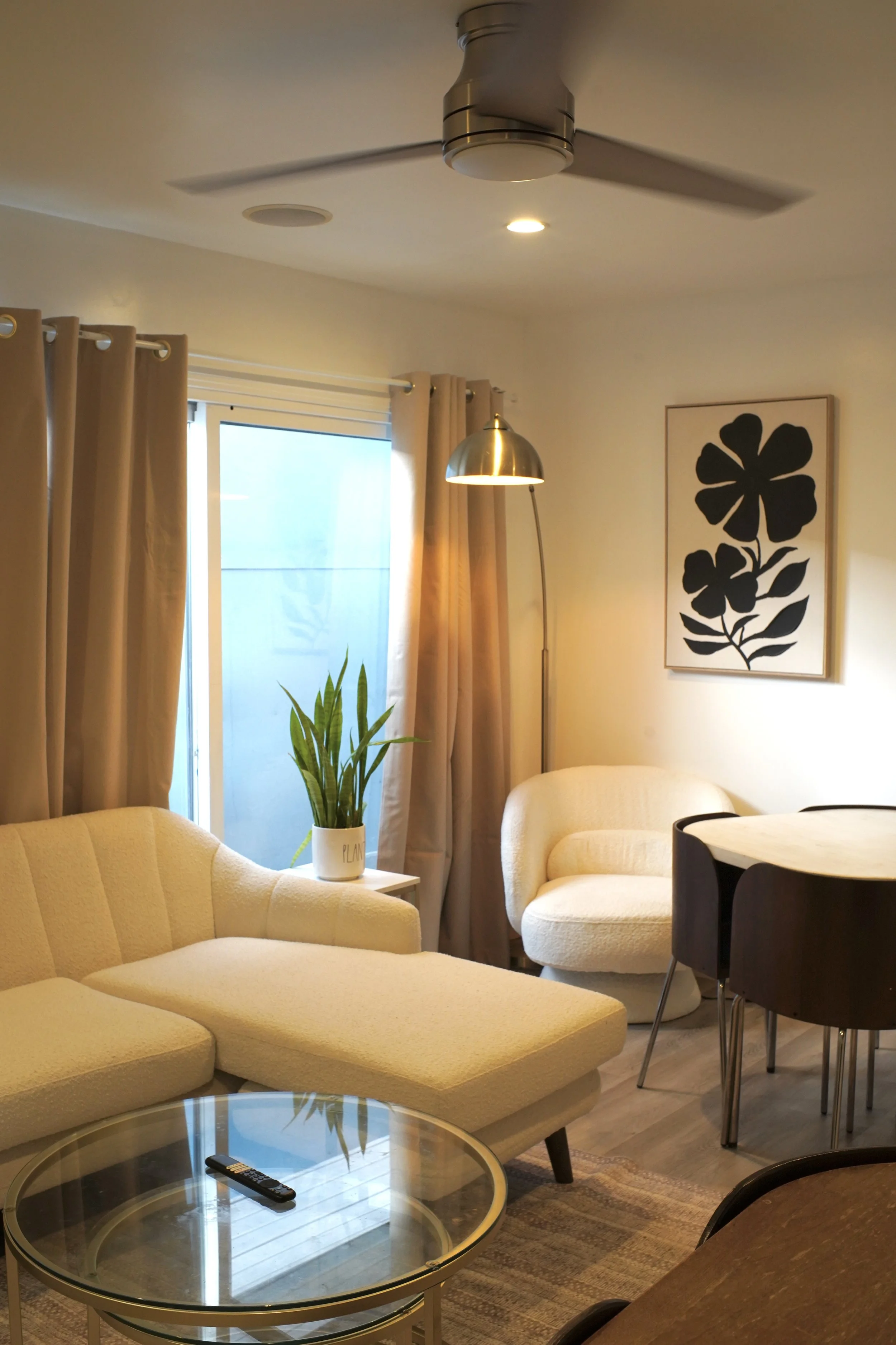 Living room with beige sofa, yellow armchair, glass coffee table with remote, potted plant labeled 'PLANT', floor lamp, artwork with black flowers, curtains, and ceiling fan.