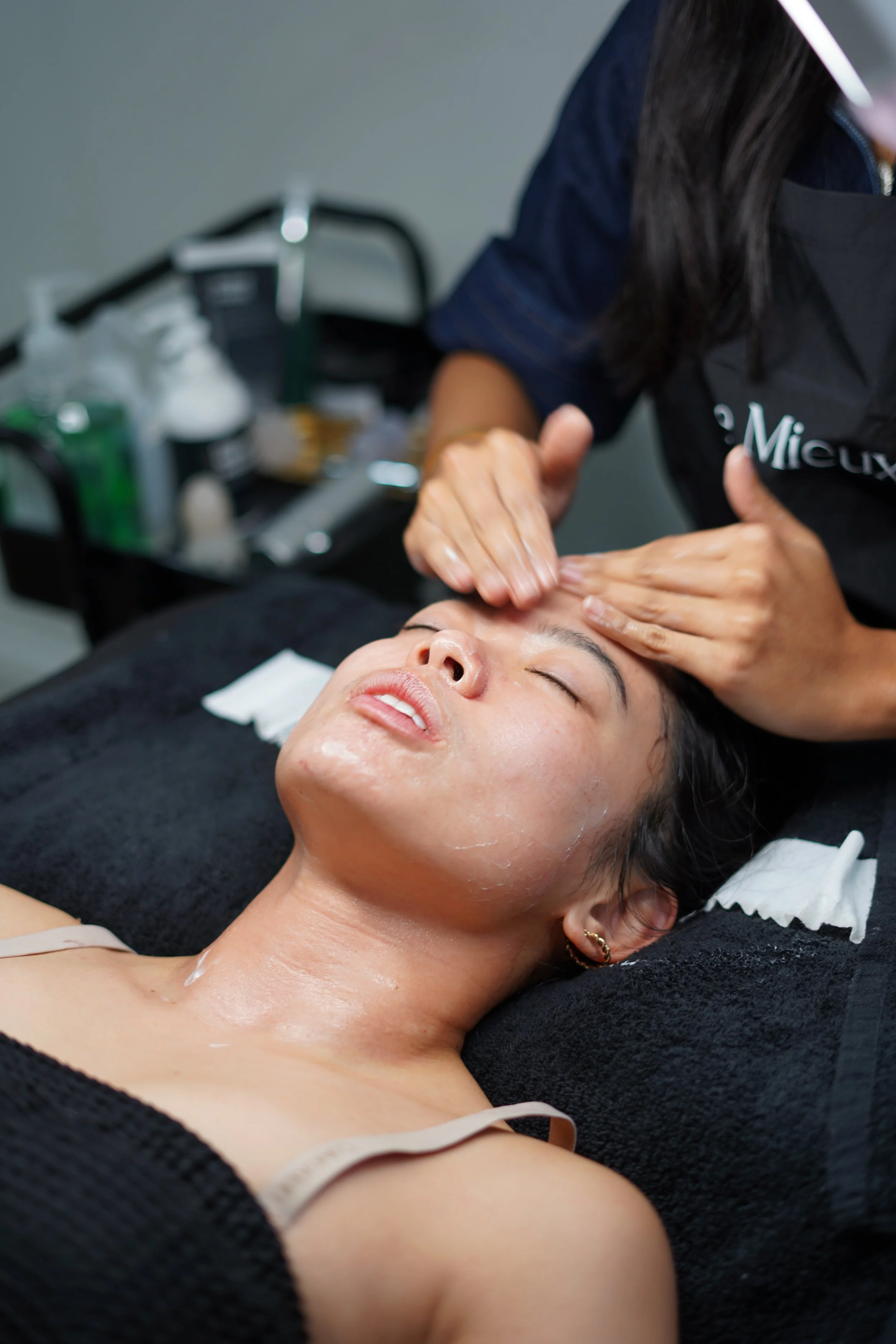 A woman is lying on a spa bed with her eyes closed while a skincare specialist applies a facial treatment. The woman is wearing a beige tank top and has jewelry in her ear. The background shows a tray with skincare products.