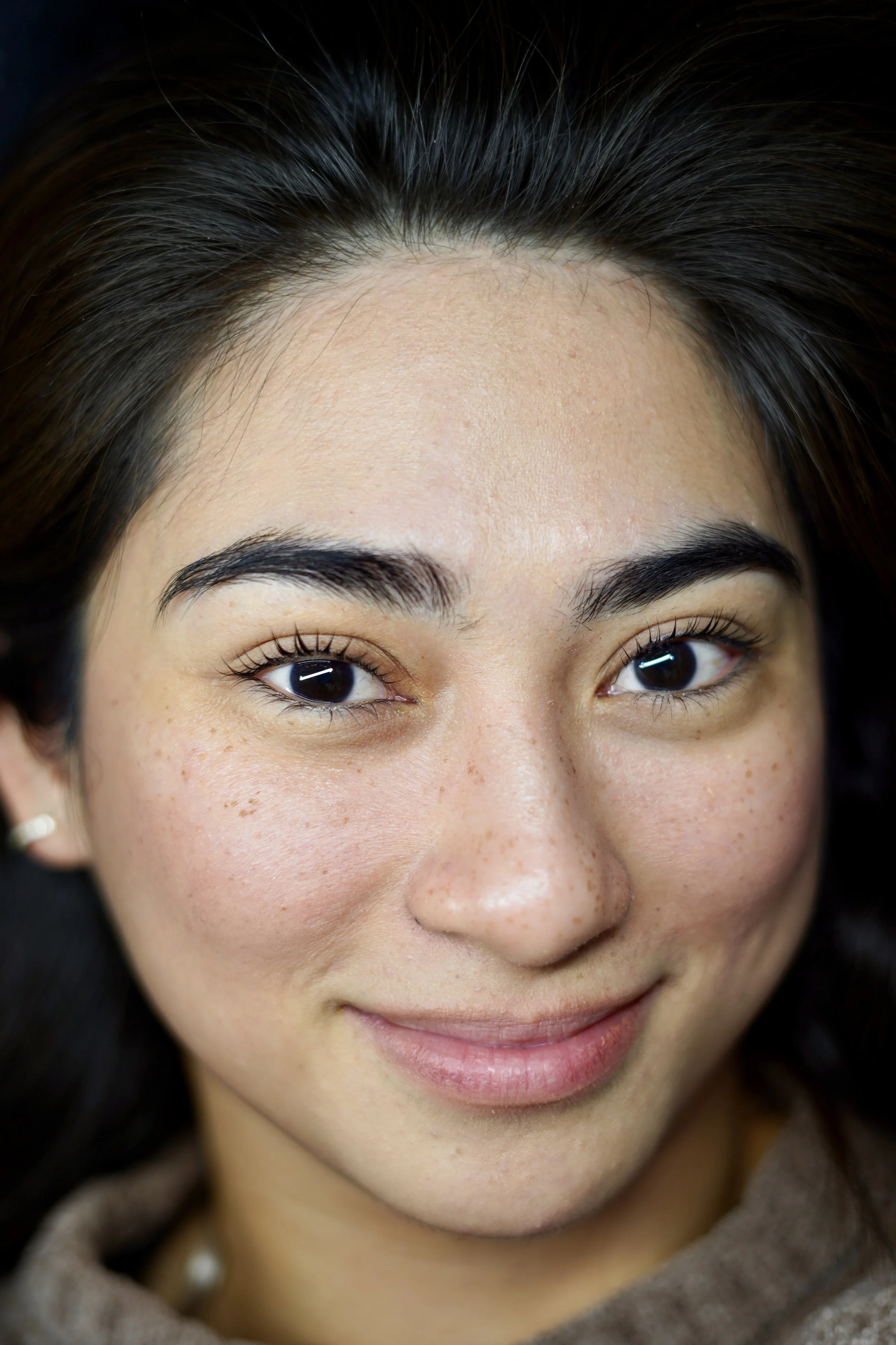 Close-up of a smiling woman's face with dark hair, freckles, and natural makeup. Korean Lash Lift results