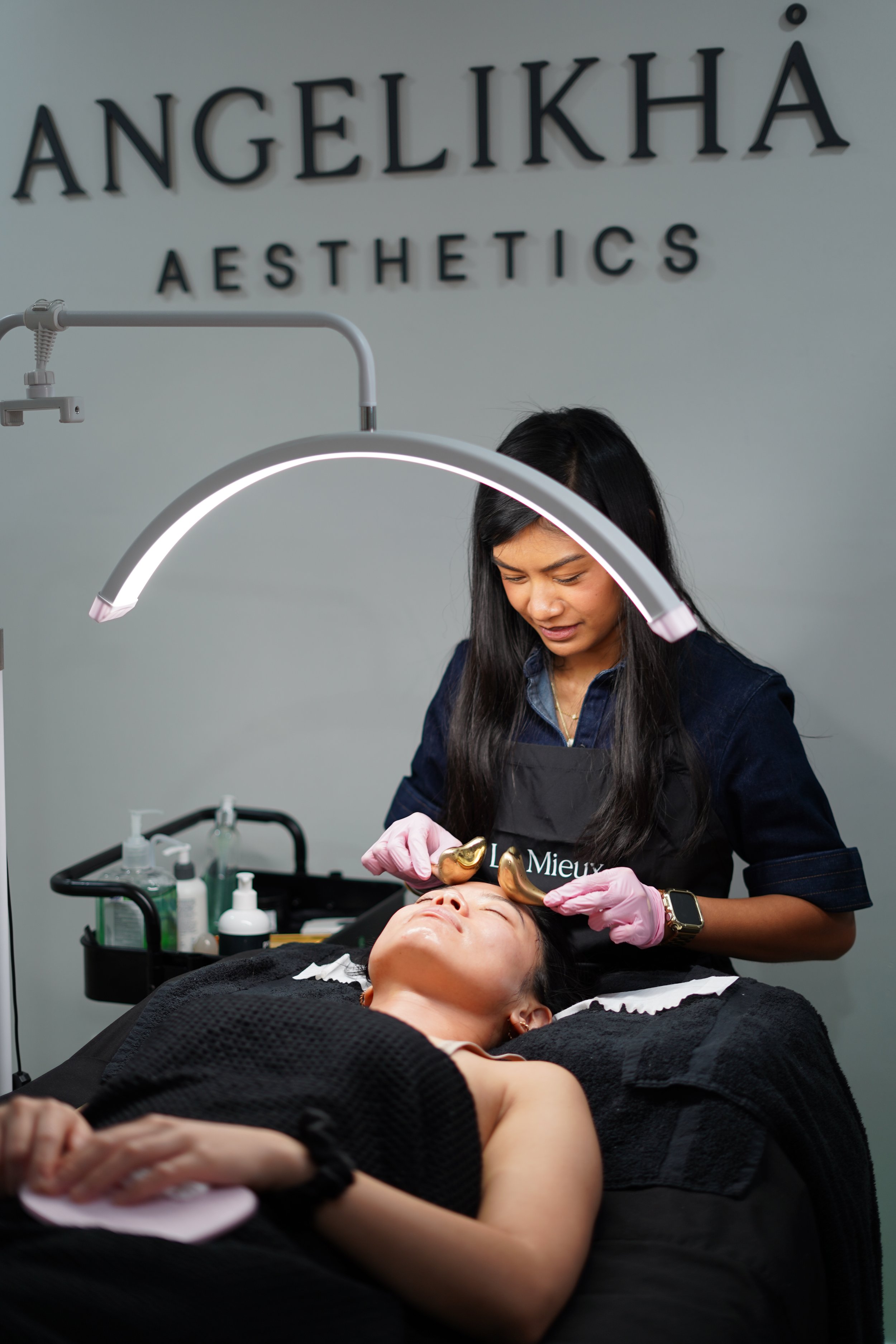 A woman receives a facial treatment at Angelikha Aesthetics, lying on a treatment bed with her eyes closed while a practitioner applies beauty tools to her face under a bright light.