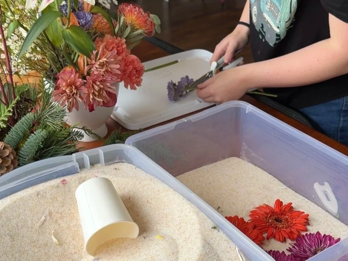 Person arranging flowers in a plastic container with silica drying beads and other colorful flowers nearby.