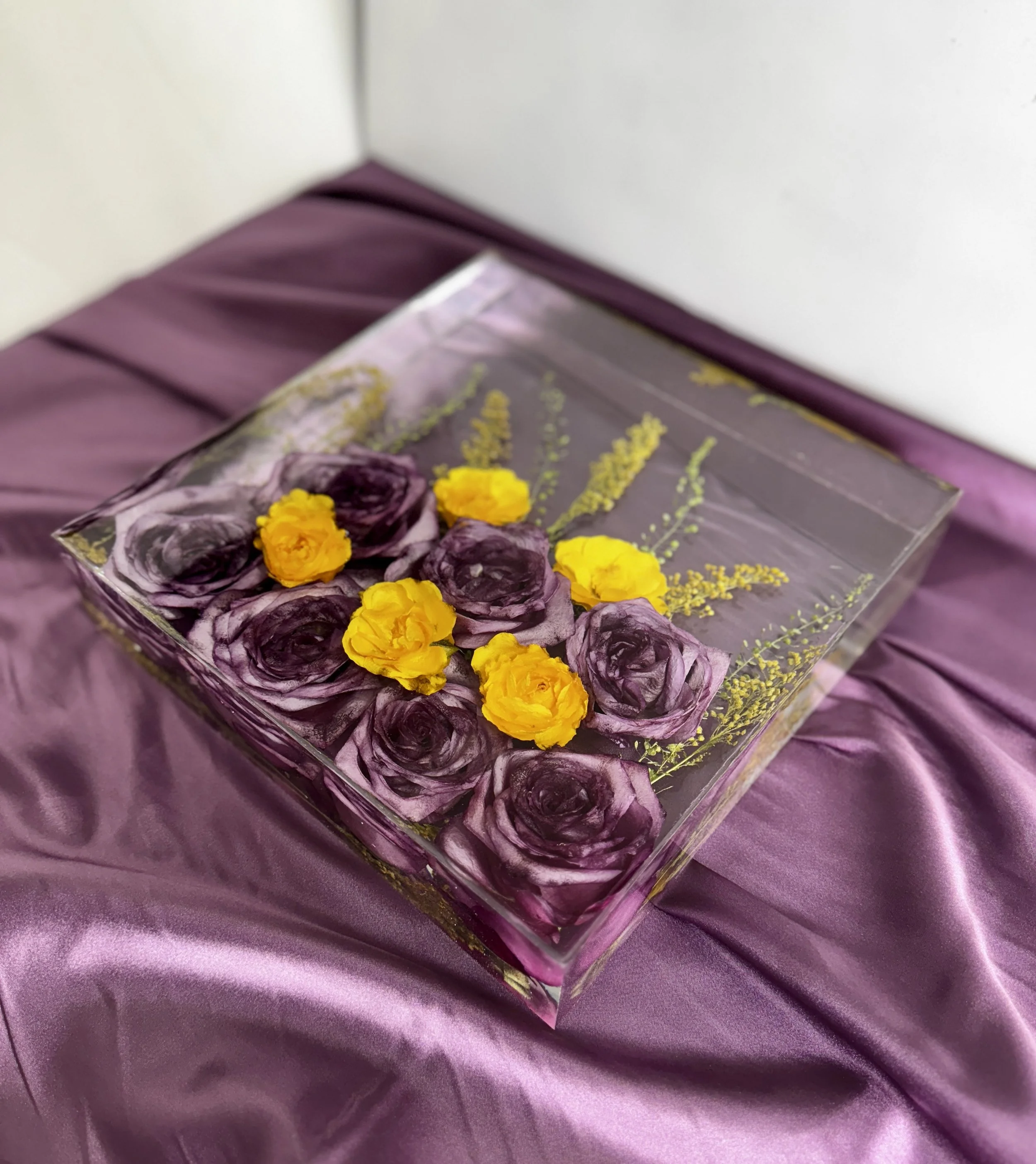 A decorative resin square with purple and yellow pressed flowers, placed on a purple satin fabric.