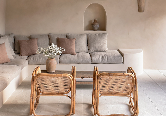 Living room with a beige sectional sofa with pillows, two rattan chairs, a wooden coffee table, a vase with white flowers, and a built-in wall niche with a decorative jar.