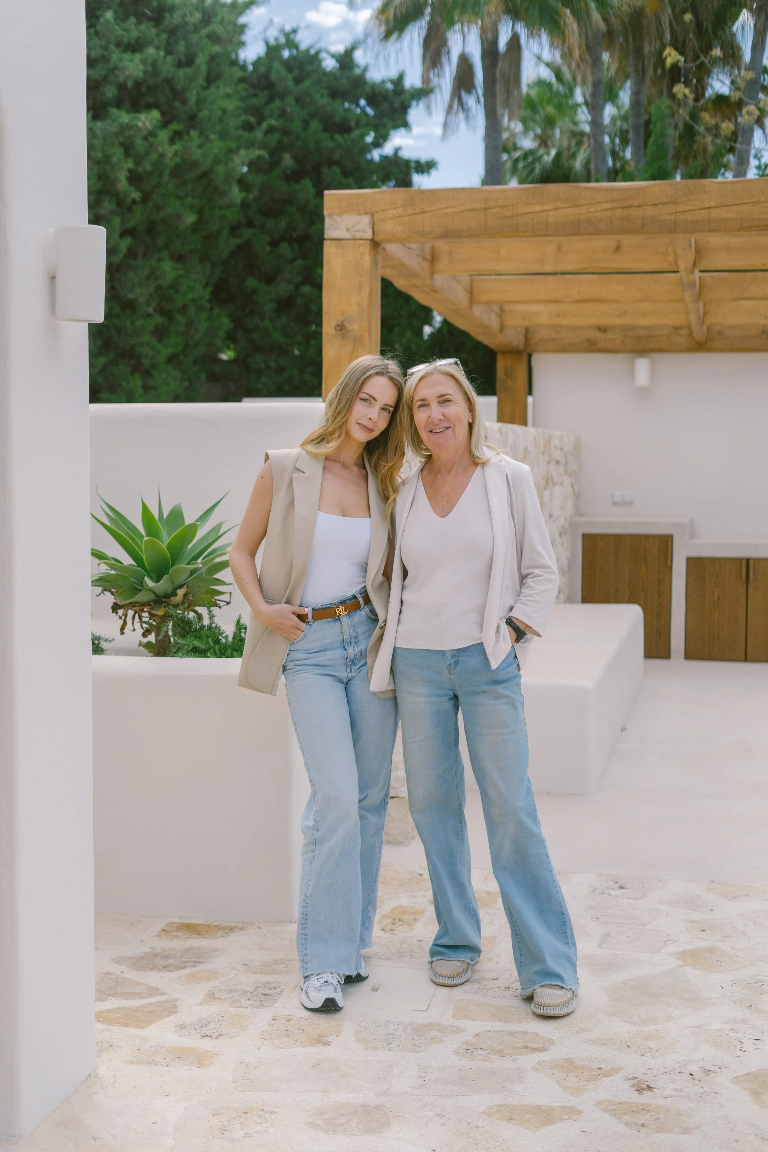 Two women, one younger and one older, standing together outdoors in a modern, minimalist setting with greenery and palm trees in the background. They are smiling and posed casually.