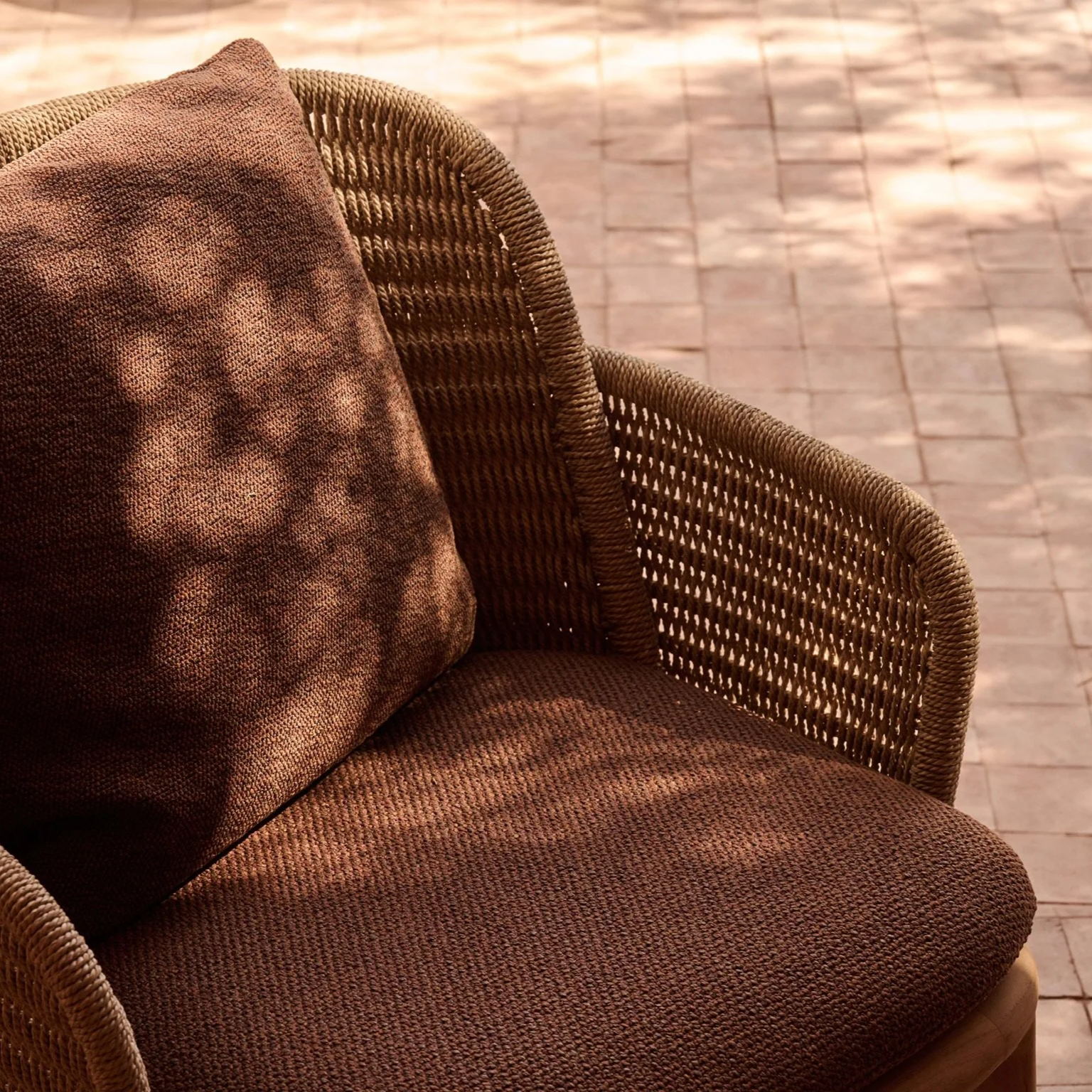 A wicker outdoor chair with a brown cushion and pillow, on a tiled brick patio.