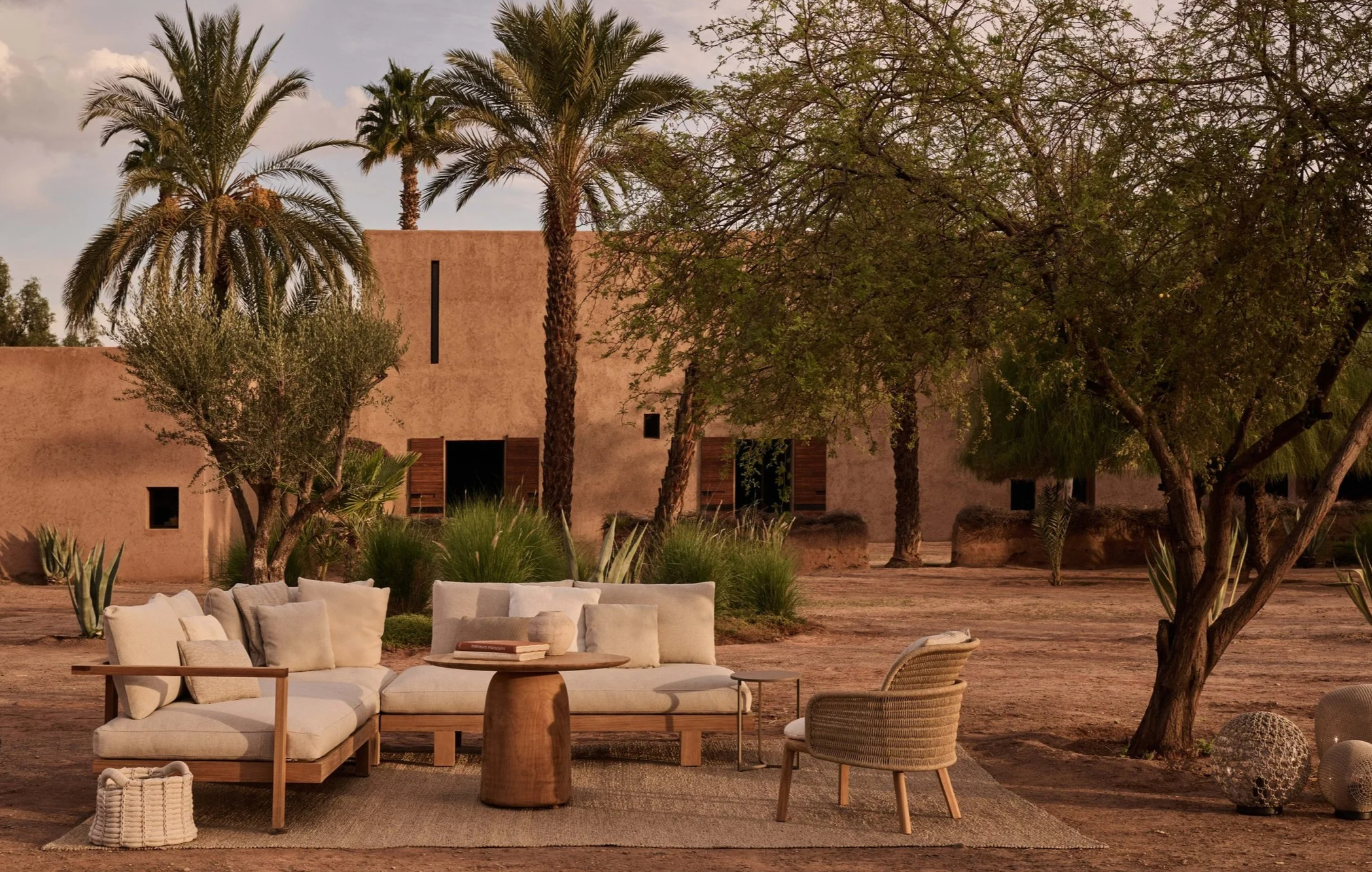 Outdoor lounge area with beige sofa, wicker chair, and wooden coffee table set on a rug in a desert landscape with palm trees, green bushes, and adobe-style building in the background during sunset.