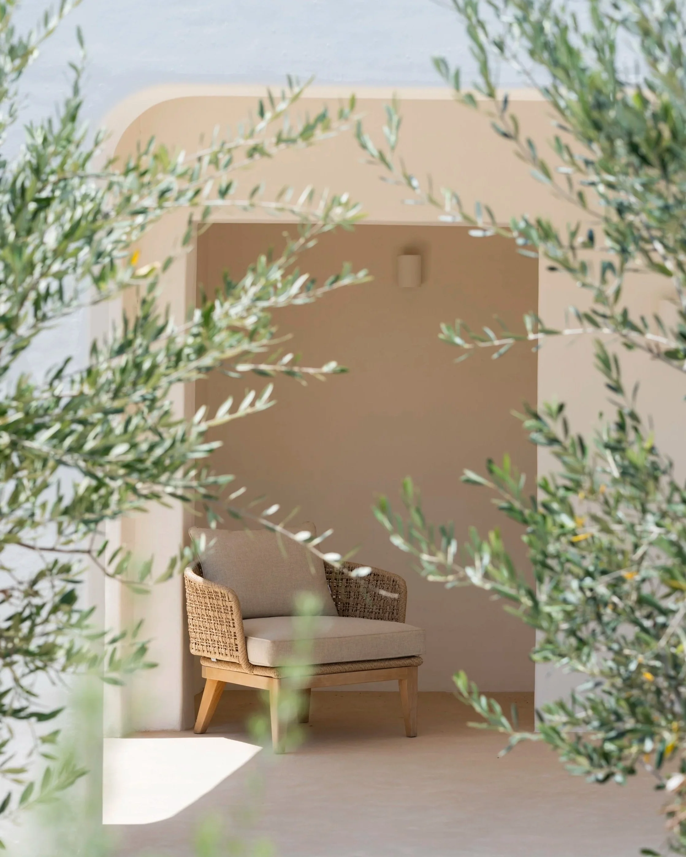 A beige armchair with wooden legs and woven sides is positioned on a light-colored porch, framed by green foliage, with a simple wall and light fixture behind it. The chair is from our interior store A concept store Moraira and is used in Casa Sabi. 