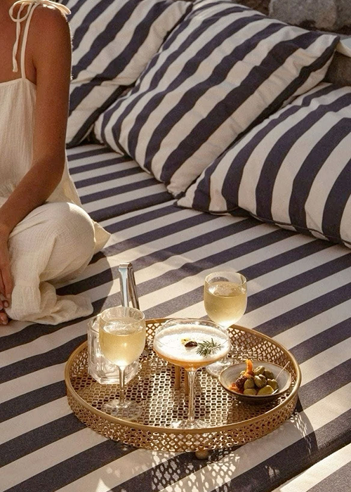A gold tray with two glasses of white wine, a cocktail, and a plate of olives and herbs on a striped bedspread in a cozy bedroom.