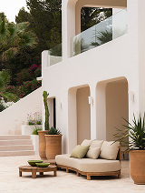 Outdoor seating area with a beige cushioned sofa, armchair, small wooden table, potted plants, and a white building with arches