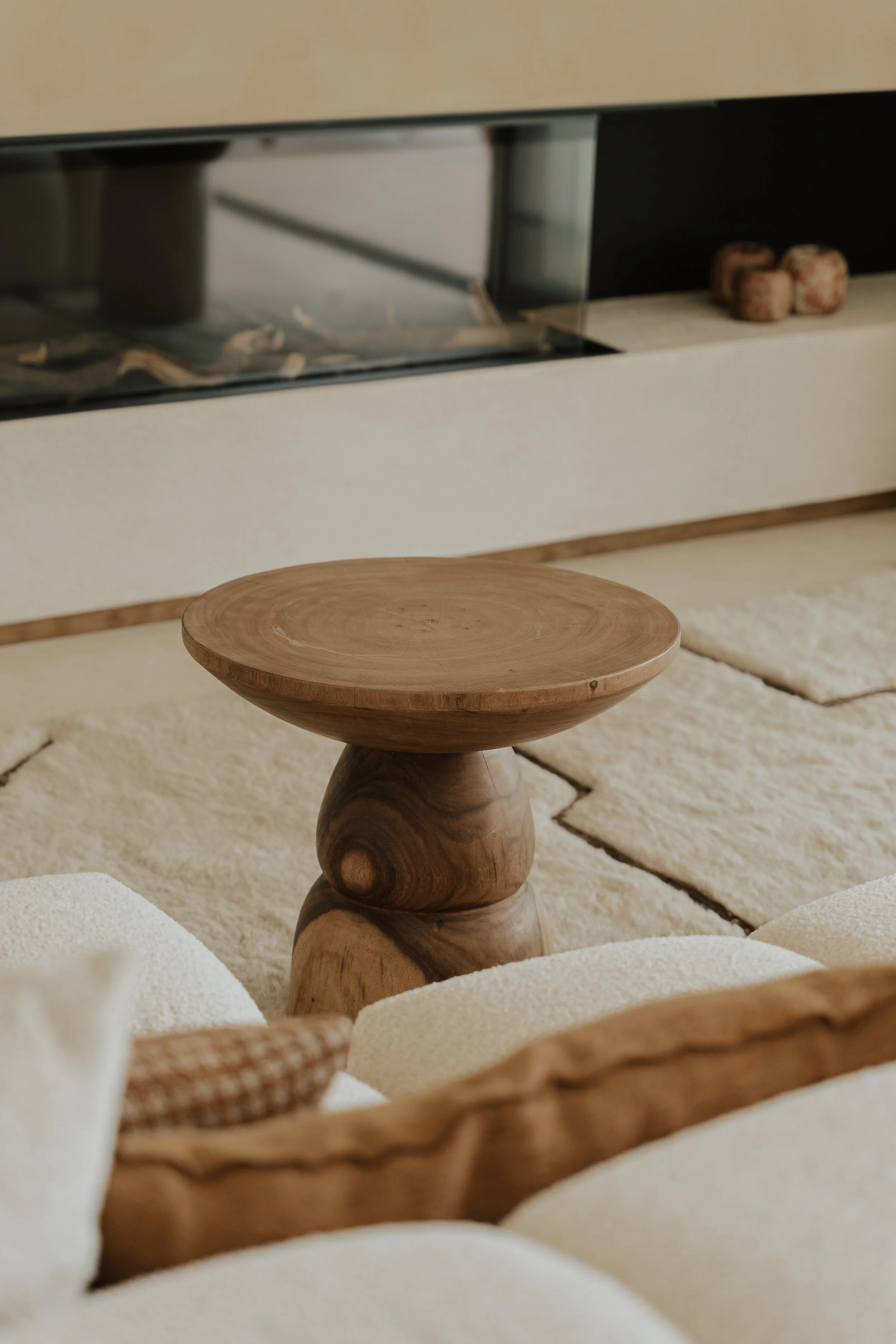 Living room thoughtfully picked out items from our interior store at the costa blanca, Moraira. Beige sofa, wooden round coffee table, and a textured cushion, against a white wall with fireplace.