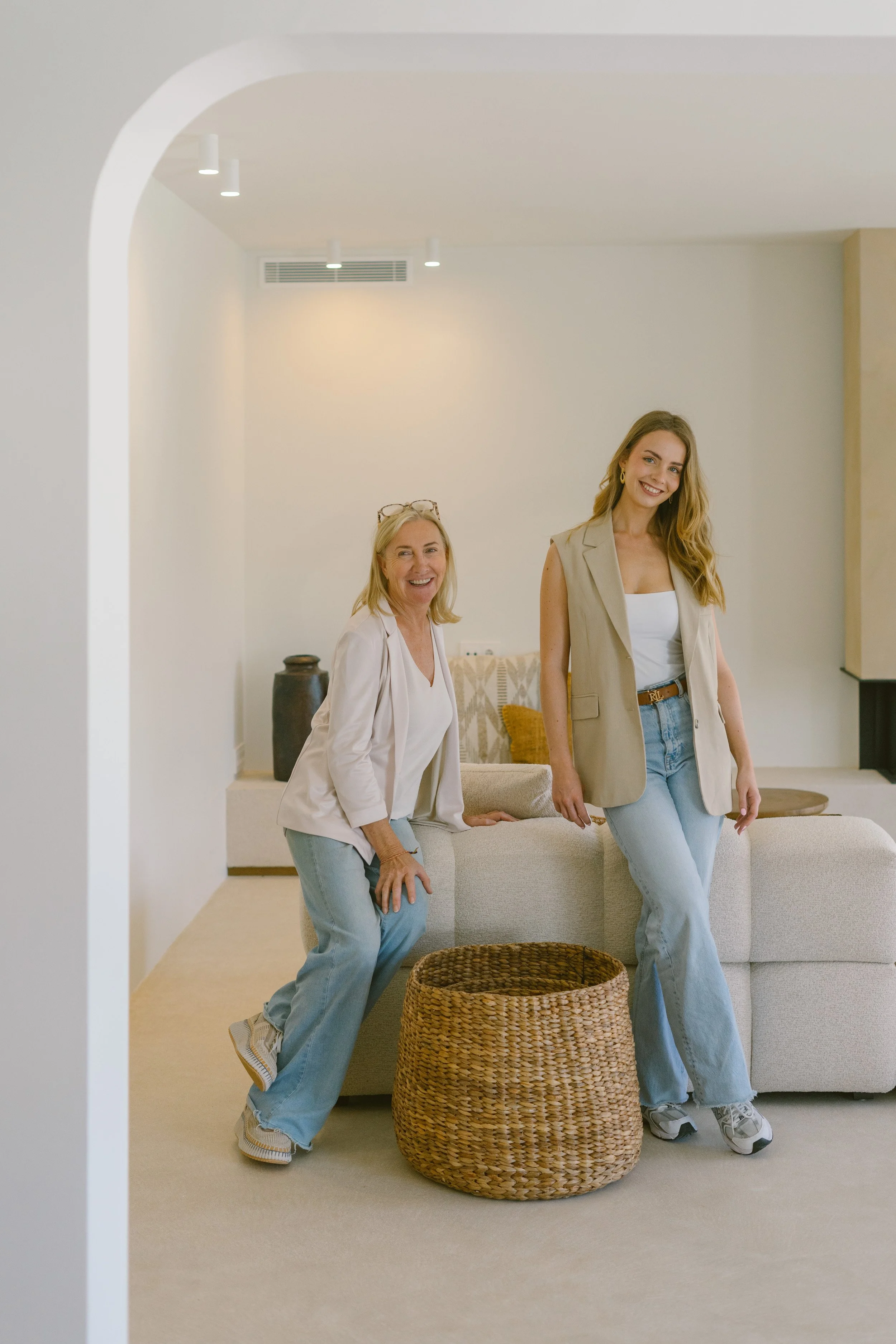 Two women, one older and one younger, smiling and posing in a modern living room with beige walls, a white sofa, wicker basket, and decorative pillows.
