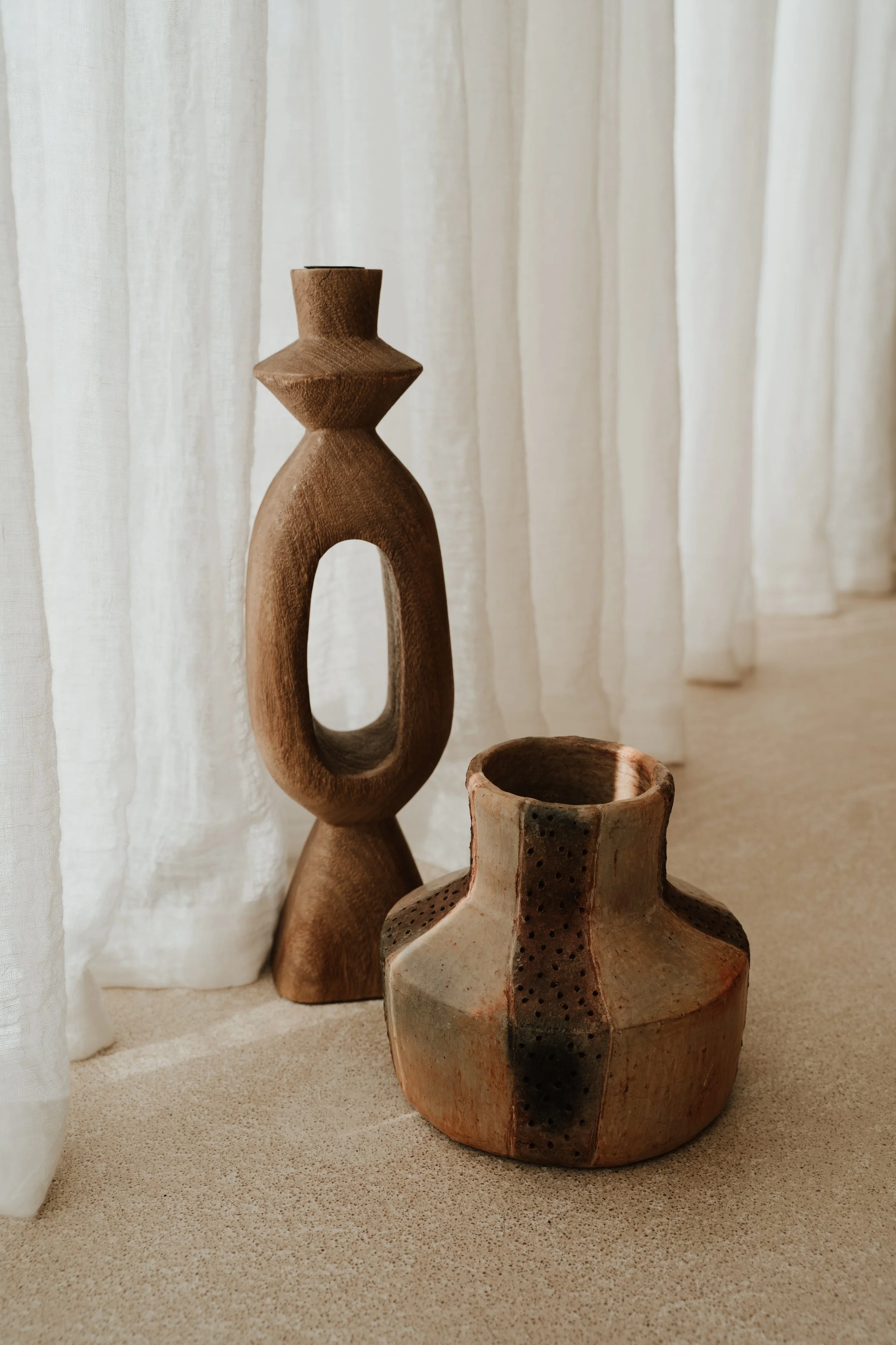 Decorative wooden sculpture and ceramic vase on a beige surface near a white linnen curtain