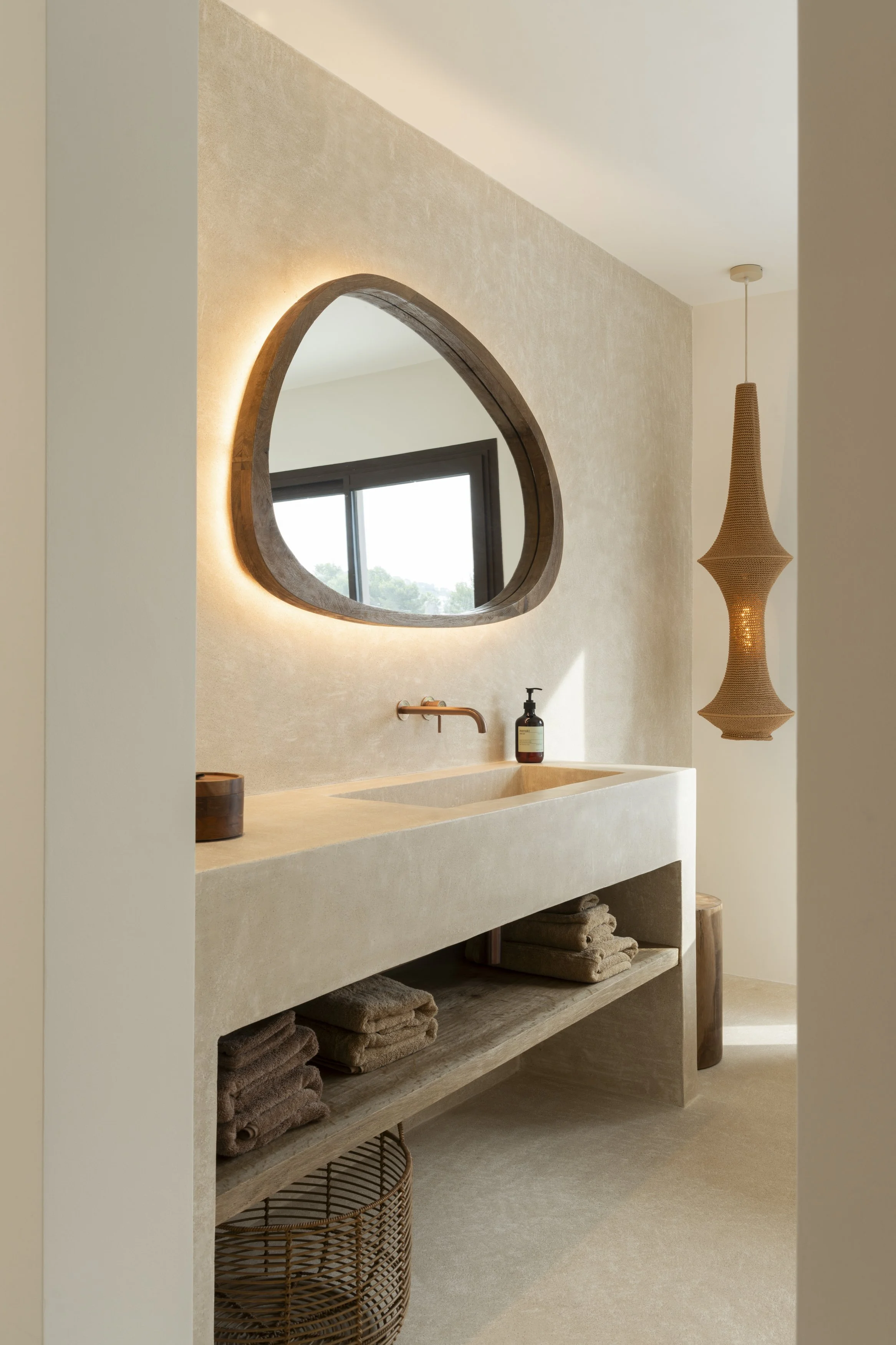 Minimalist bathroom with a cream-colored stone sink, round copper faucet, wooden mirrors, and beige towels. A woven pendant light hangs from the ceiling.