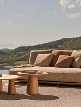 Outdoor seating area with a beige sofa and wooden coffee tables near a lake, surrounded by greenery and hills.