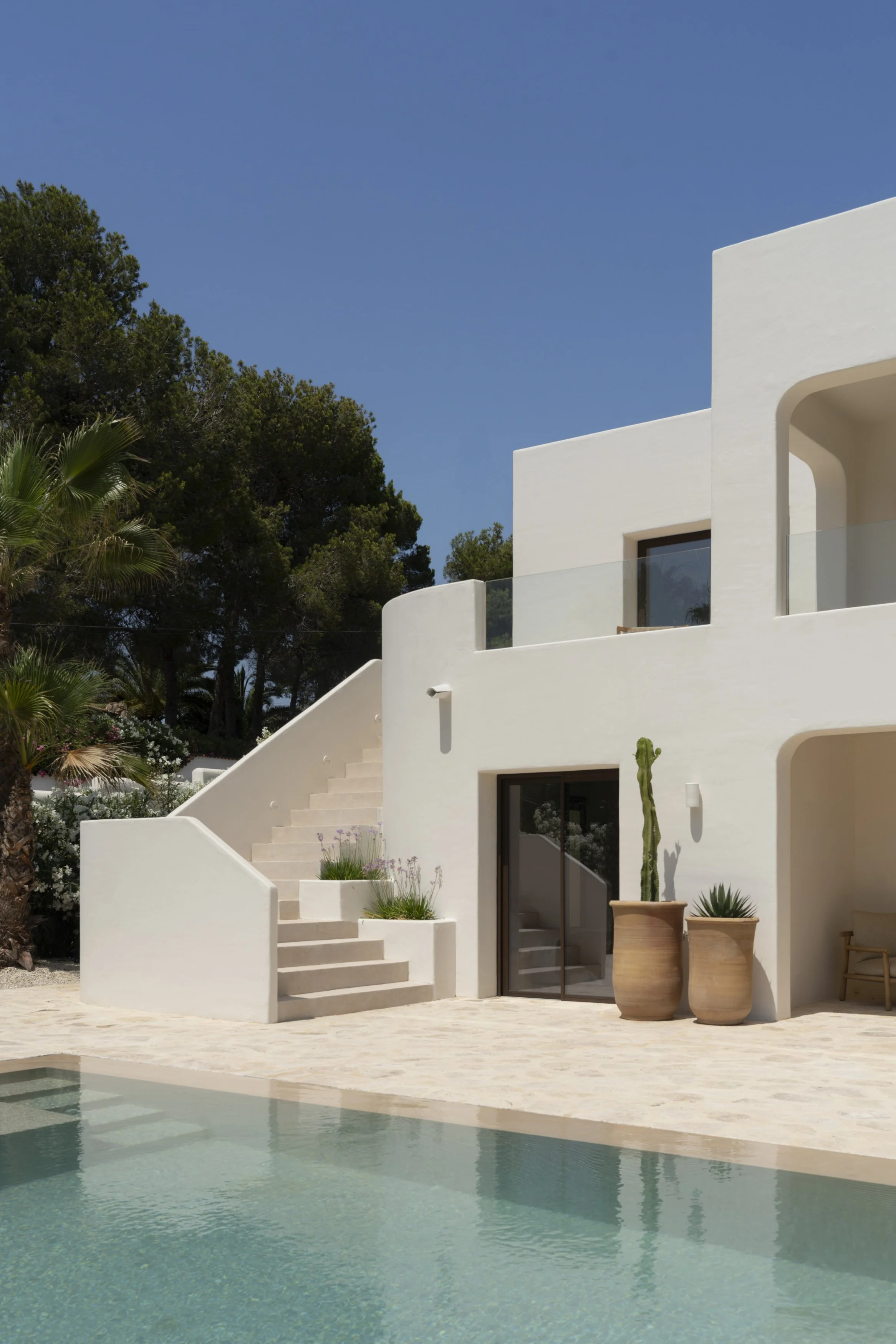 A modern white house with outdoor stairs, large glass doors, and balcony, next to a swimming pool, with potted plants and a clear blue sky.